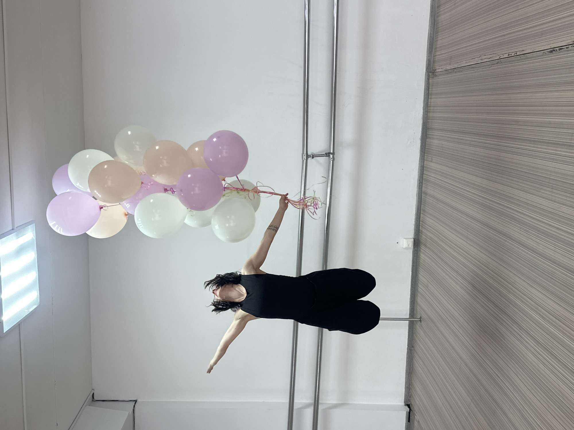 A woman in black clothing is suspended horizontally in mid-air, holding a bunch of pastel-colored balloons. She is attached to a metal pole via a harness. The location is a room with white walls and a wooden panel wall. There are two metal poles running vertically along the wall. The lighting is bright and artificial. The scene is indoors. The city of Votkinsk, Russia is not discernable.