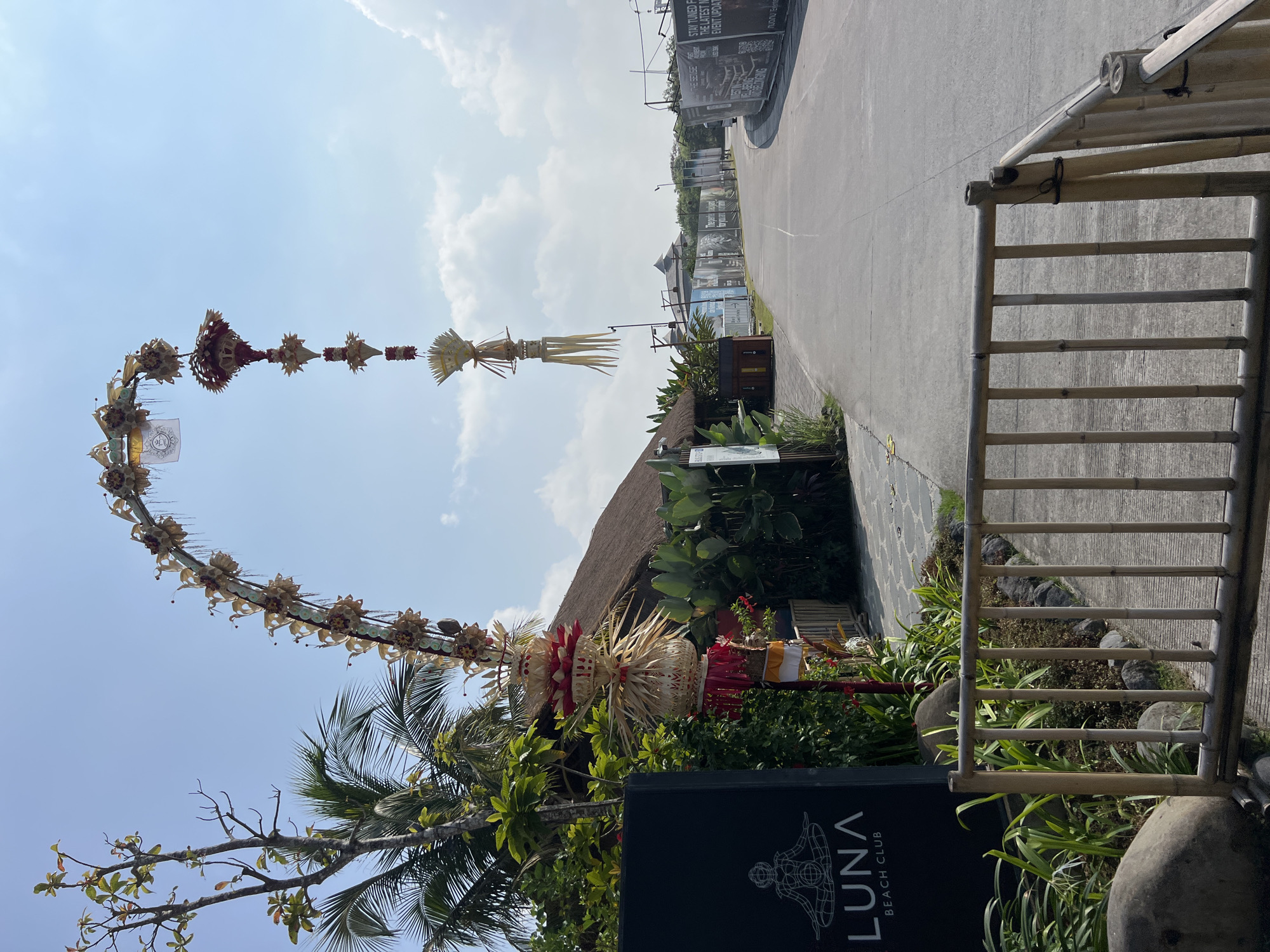 The image depicts an outdoor scene with a prominent arched bamboo structure adorned with traditional Balinese decorations, possibly a "penjor" or a similar festive archway. This archway spans over a paved pathway or road. To the left, a black sign with white lettering reads "LUNA BEACH CLUB," suggesting the location is a beach club or a venue with that name. Behind the sign and the archway are lush green vegetation, palm trees, and a building with a thatched roof, characteristic of tropical architecture. The sky is clear with scattered clouds. In the background, a road extends into the distance, lined with fences and what appear to be construction barriers or advertisements. The overall impression is of a welcoming entrance to a resort or entertainment venue in a tropical setting.