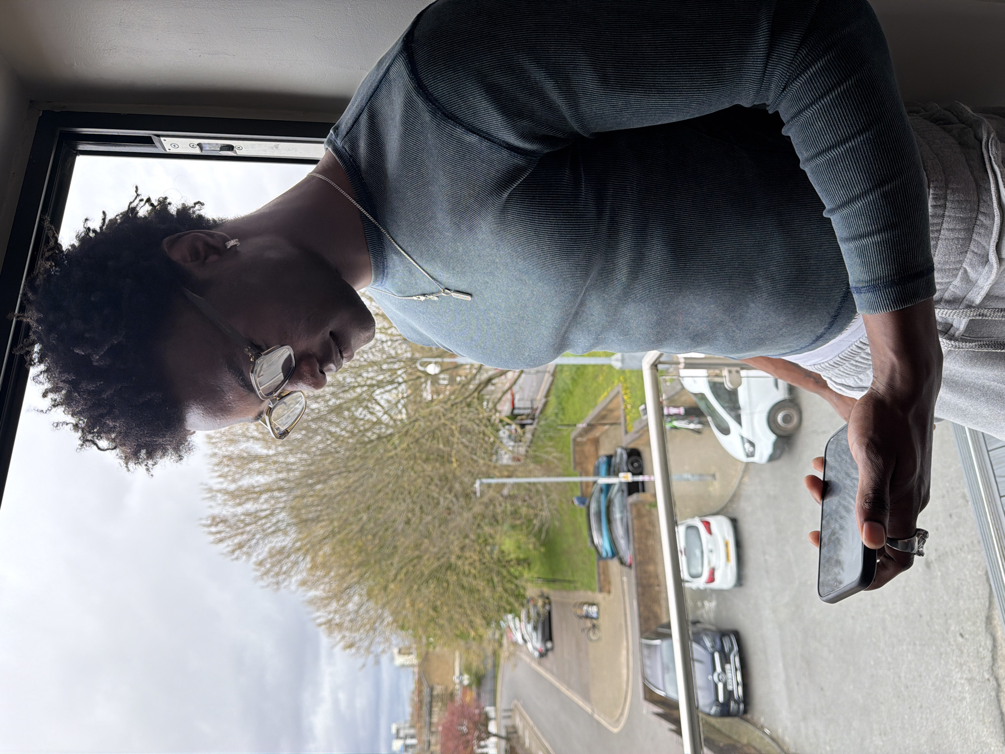 A person stands at a window, angled towards the outside. The individual has dark curly hair and wears glasses. They are dressed in a long-sleeved, dark teal ribbed shirt and light gray sweatpants. A silver necklace is visible. The person is holding a smartphone in their right hand, looking down at it. On the hand holding the phone, there is a prominent silver ring.
Outside the window is a view of a street in what appears to be a residential area. Several cars are parked along the side of the road. In the background, buildings and trees are visible. The sky is overcast.
Determining the specific location within London would require further visual clues such as street signs, architectural styles, or landmarks.
