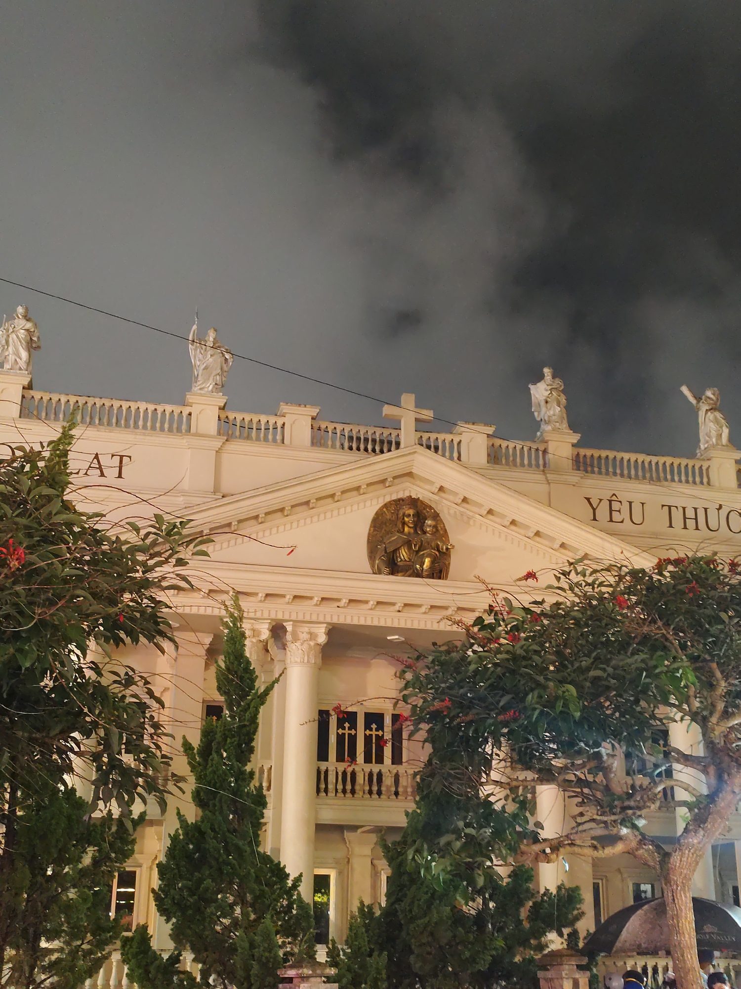 The image captures a grand, classically designed building illuminated at night under a dark, cloudy sky. The structure features a prominent pediment with a golden bas-relief sculpture depicting religious figures, flanked by large white columns. Balustrades line the roof, adorned with several statues. A white cross stands centrally on the roofline.
On the left side of the building's upper facade, the partial text "LAT" is visible, likely indicating "DALAT," while on the right, the Vietnamese words "YÊU THƯƠNG," meaning "Love," are displayed. Statues of figures in classical attire, some holding objects like a staff or spear, stand along the roofline. A power line cuts across the dark sky above the building.
The foreground is filled with lush greenery, including tall coniferous trees and leafy shrubs bearing small red flowers, partially obscuring the lower levels of the building. The artificial lighting highlights the architectural details and statues against the dark, overcast sky, creating a dramatic nighttime scene. Hints of people can be seen at the very bottom right corner, suggesting a public area. The setting is clearly Da Lat, Vietnam, as indicated by the visible text and context.