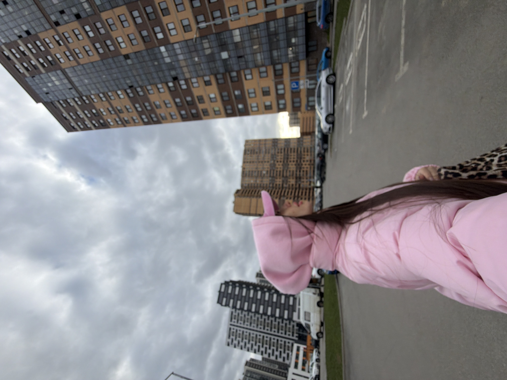 A casual, full-body shot captures a person standing in an urban environment under an overcast sky. The main subject, positioned on the right side of the frame, is seen from the mid-torso up, wearing a light pink puffer jacket. Long, dark hair falls over their back, and a hint of a leopard print pattern is visible on their left shoulder, possibly from a top or bag. Their head is tilted slightly upwards and to the left.
The background is dominated by several modern, multi-story residential buildings. A large building on the left features a distinctive brown and gray facade with numerous rectangular windows. Further back, another textured brown building is visible, alongside other contemporary structures. The ground consists of an asphalt parking lot with white painted markings. Several cars are parked along the right side of the lot, including a light-colored sedan and a blue car, with a white van also visible in the distance. A blue parking sign with a white 'P' symbol is discernible near the parked vehicles. The sky is heavily clouded, indicating a cloudy daytime setting. The scene depicts a typical residential area within a city, consistent with the provided context of Saint Petersburg, Russia.