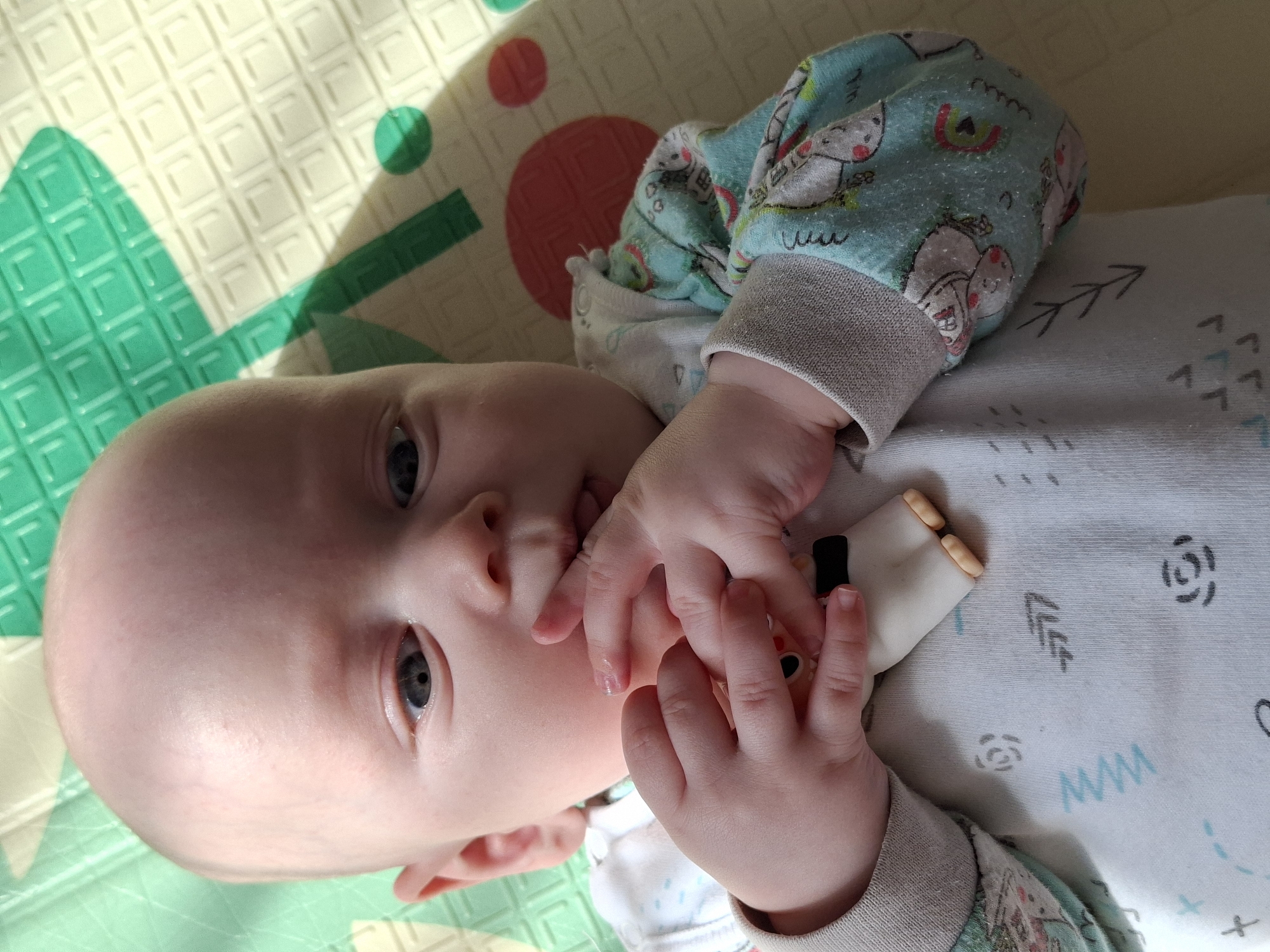 A baby is lying on a play mat. The baby is wearing a light blue and white patterned onesie with gray cuffs. The baby's hands are near their face. One hand is holding a toy. The play mat is multi-colored with geometric shapes. Sunlight is shining on the mat. The location is unknown.