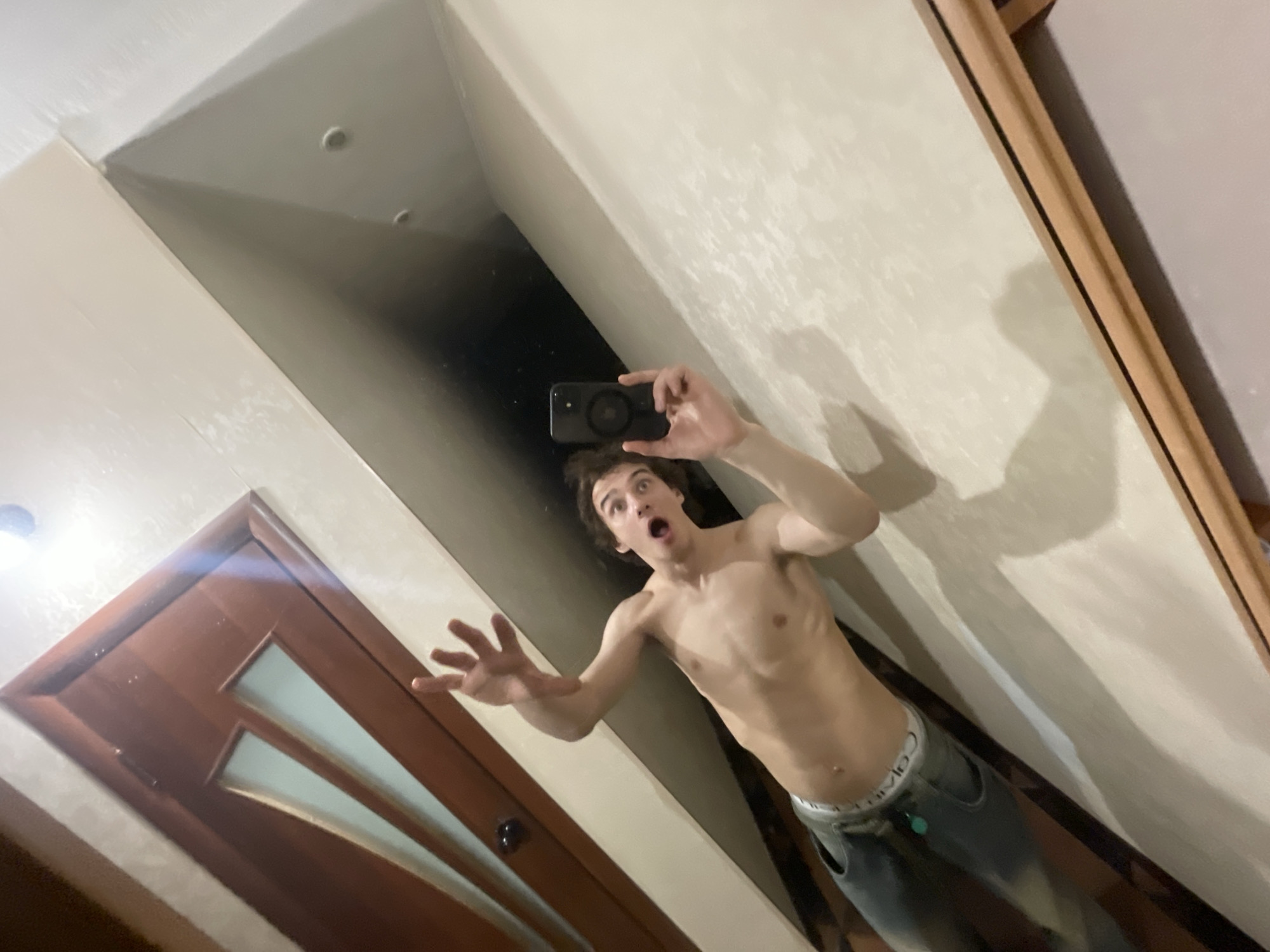 A young man is taking a selfie in a mirror. He is shirtless and wearing jeans with a white waistband. His expression is one of surprise or fear. He is holding a black smartphone in his right hand, taking the photo. The reflection shows a hallway. The location is an apartment and the city is presumed to be Krasnoyarsk, Russia. A brown door with translucent panes is visible on the left side of the reflection. The walls are light-colored with textured paint. A wooden frame outlines the mirror. The lighting appears to be from indoor sources.