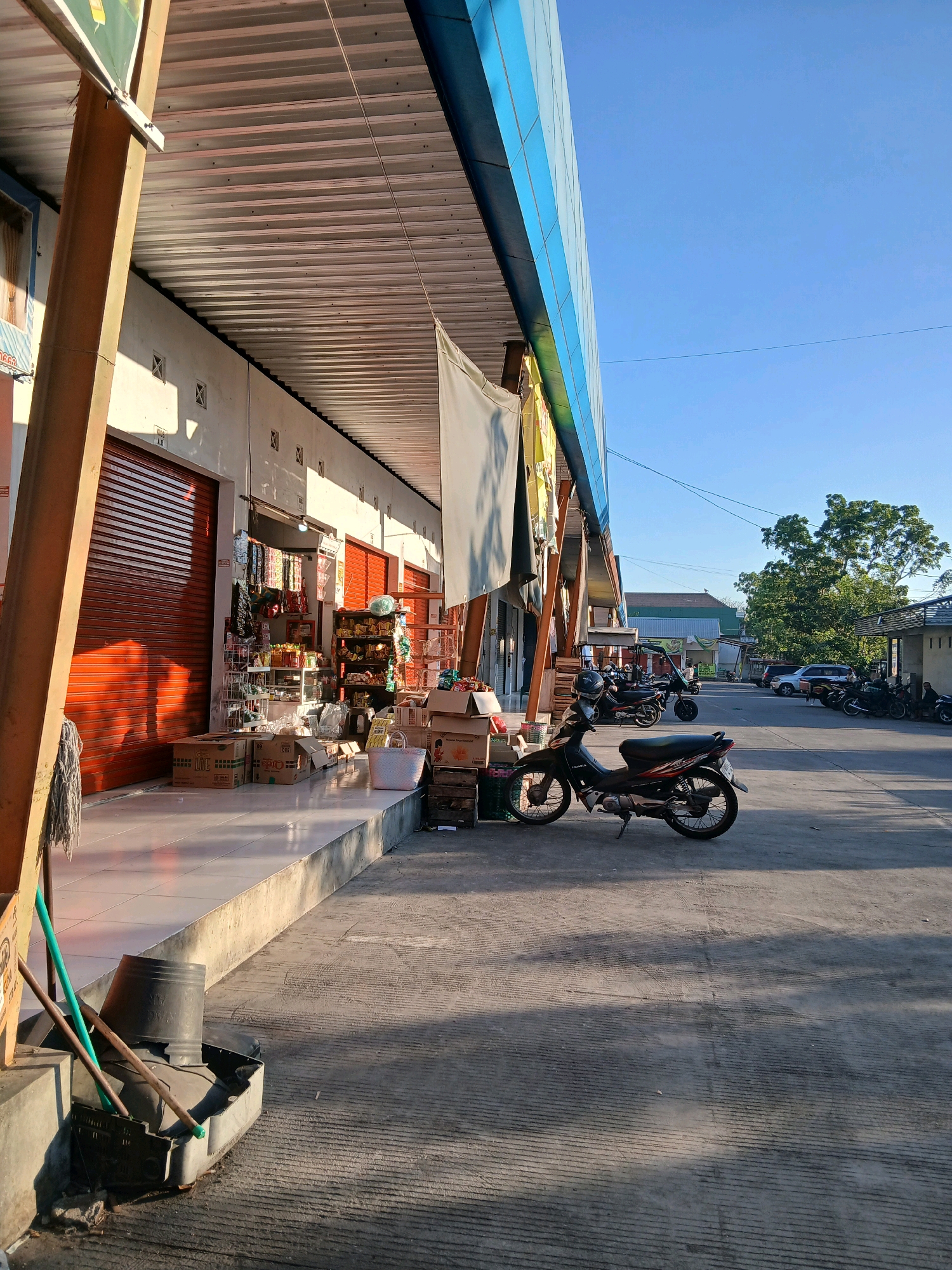 The image depicts a street scene in front of a row of shops in Loram Wetan, Indonesia. The shops have corrugated metal roll-up doors, some of which are red. Merchandise, including bags of goods and boxes, is displayed on shelves and on the ground outside some of the shops. A parked motorcycle is in the foreground, and other motorcycles and cars are visible further down the street. The sky is clear and blue, and the sun casts long shadows. The overall impression is one of a typical market or commercial area in a warm climate.