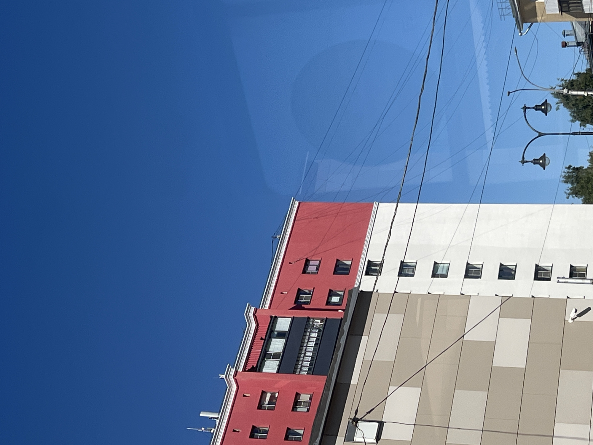 The image captures an urban scene with a bright blue sky. In the foreground, a multi-story building is visible. The upper section of the building is painted bright red, with a white trim and several windows. Below the red section, a portion of the building is white, also featuring multiple windows. Further down, a section of the building is clad in beige panels arranged in a grid pattern. Another building, partially visible on the right, appears to have a yellow facade and a sloped roof. Several streetlights with black lamps are positioned in front of the buildings. Electrical wires crisscross the sky. The scene is set in an urban environment, likely a city.