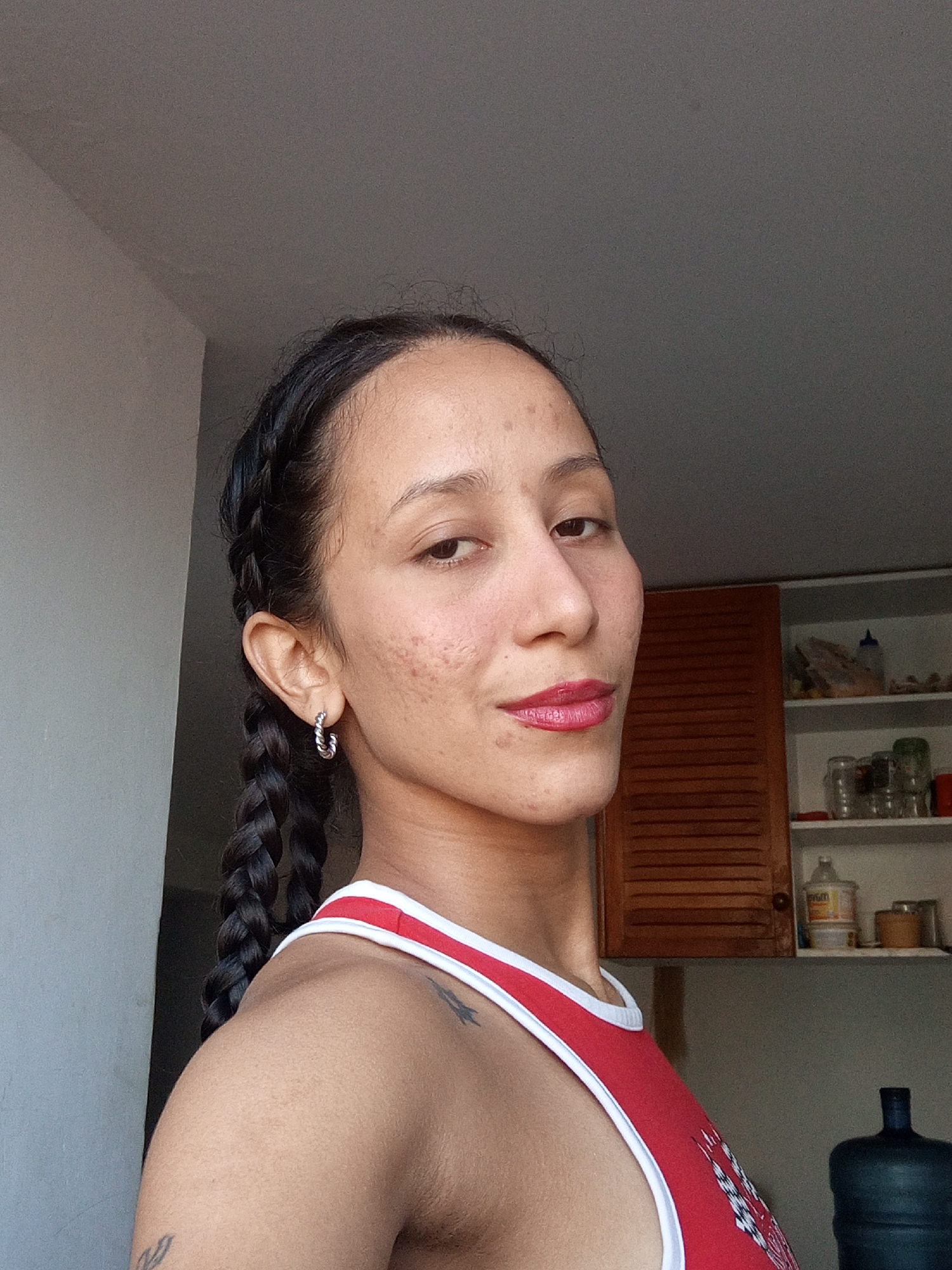The media file is a portrait-oriented, eye-level, medium shot of a young woman with dark hair styled in two braids. She is wearing a red tank top with white trim. Her skin has some blemishes. She is looking towards the camera with a slight smile. Her makeup includes pink lipstick. She is wearing a silver hoop earring in her left ear. A tattoo is visible on her left shoulder.
In the background, there is a wooden cabinet with horizontal slats and a set of shelves filled with various items, including jars and bottles. To the right of the cabinet, a large dark blue water cooler bottle is visible. The lighting suggests it is daytime, possibly indoors with natural light from a window, given the shadows and the angle of light on her face. The scene appears to be domestic, possibly a kitchen or living area. The woman's expression is relaxed and friendly. No text is visible in the image. The provided location context is Porlamar, Venezuela.
