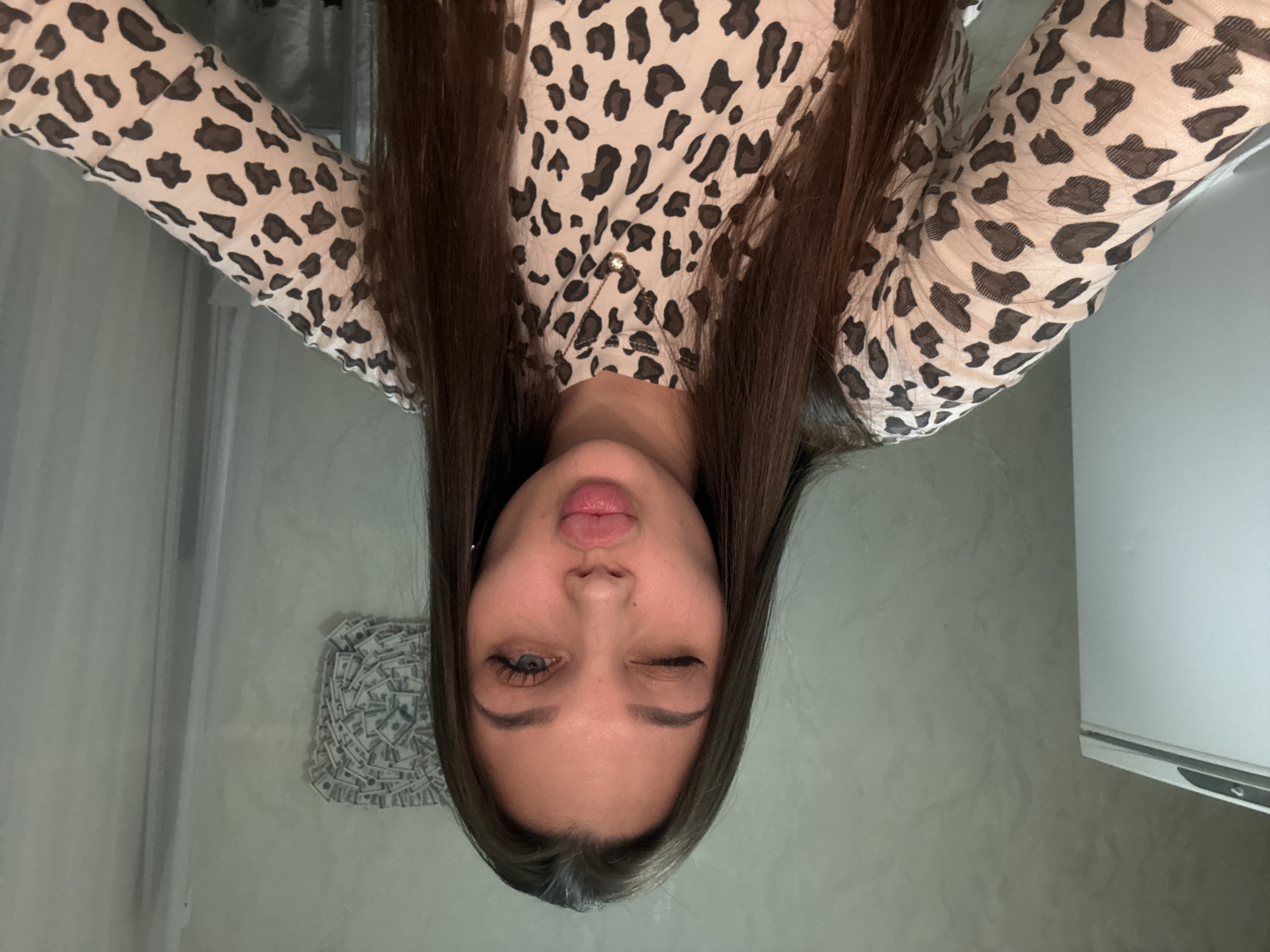 A young woman with long brown hair is upside-down in the image. She is wearing a long-sleeved, leopard print shirt. She is winking with her right eye and puckering her lips. In the background is a light gray wall and a small picture or decoration made to look like a pile of US dollar bills. Part of a white appliance is visible to the right. The specific location in the city of Chaikovsky, Russia is not discernible from the image.