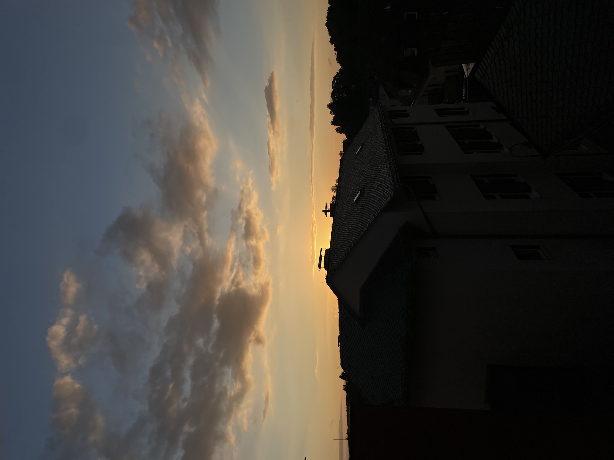 This image depicts a sunset over the city of Waldbröl, Germany. The sky is a gradient of blues and oranges, with scattered clouds illuminated by the setting sun. Silhouetted against the sky are several buildings, including a prominent multi-story structure with a tiled roof and several dormer windows. Another building to the left, in darker silhouette, has a stark, unadorned facade. Trees and foliage form a dark band along the horizon, further emphasizing the contrast with the brightly lit sky. The scene is devoid of any visible human activity.