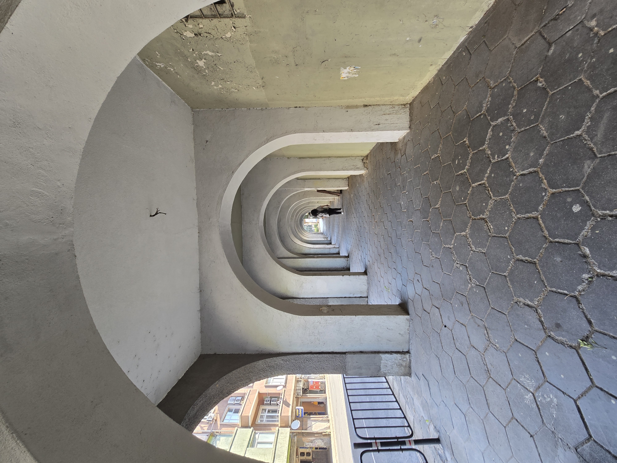 The image captures an architectural walkway formed by a series of repeating arches, creating a tunnel-like perspective. The arches are constructed from gray concrete or plaster, showing some signs of aging and discoloration. The walkway's floor is paved with dark gray hexagonal tiles.
At the far end of the walkway, a person can be observed standing, partially obscured by distance. The area outside the arches reveals buildings of moderate height. The weather seems to be fairly neutral, with no heavy shadows. The scene appears to be taken in an urban environment.