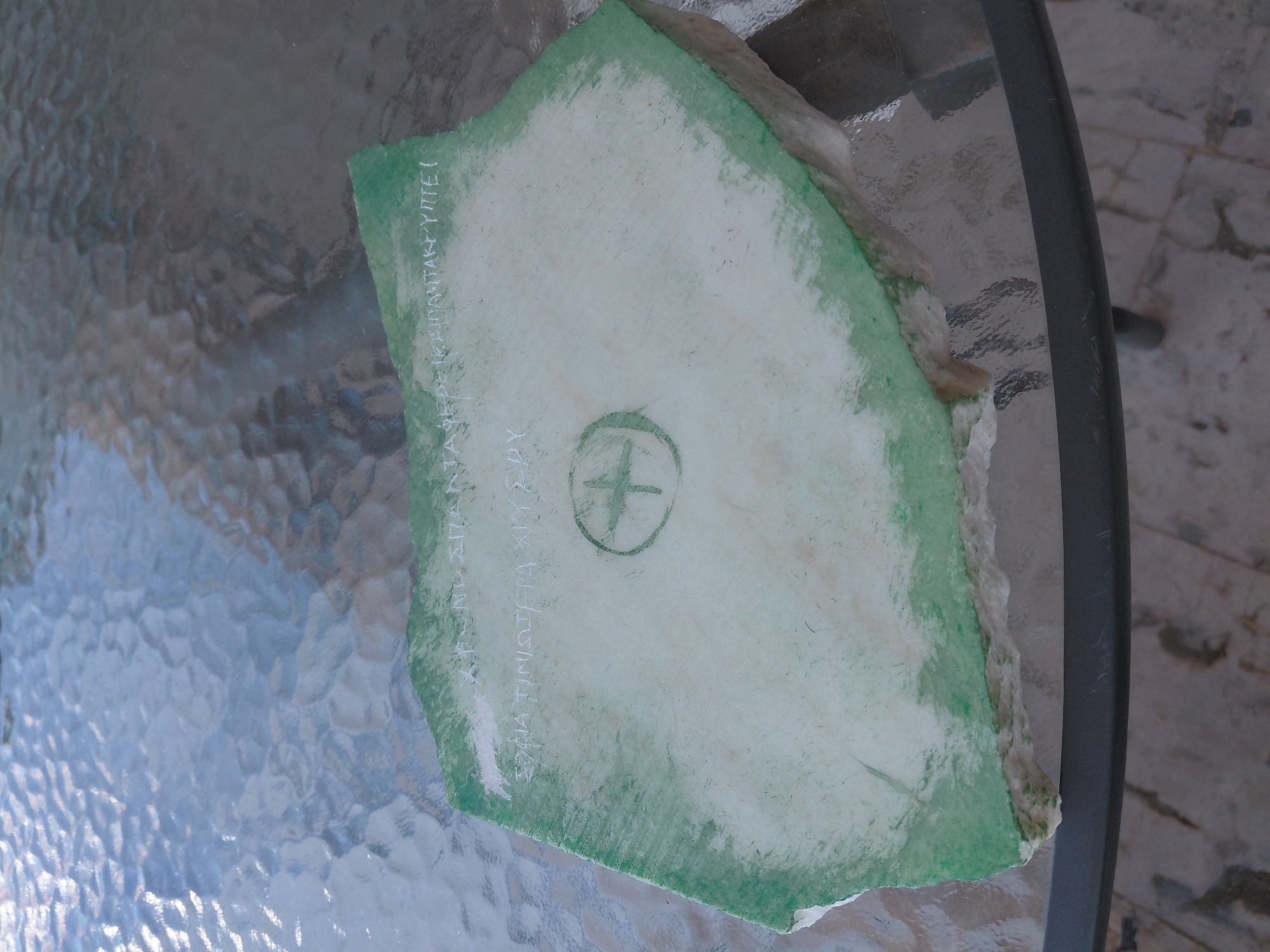 A flat, irregular-shaped piece of marble or stone, approximately 2-3 feet in length, rests on a textured glass tabletop. The stone is partially covered with a white or light-colored material, possibly plaster or paint, with a green border painted around its perimeter. Inscribed in white on the light surface are Greek characters and a circular symbol with a cross in the center, resembling an emblem. The stone appears to be located outdoors on a table with a metal frame, suggesting it might be a decorative item or a fragment of a larger structure. The background shows a tiled or paved surface and hints of natural light. The precise location within the Municipal Unit of Aegio, Greece, is not discernible from the image.