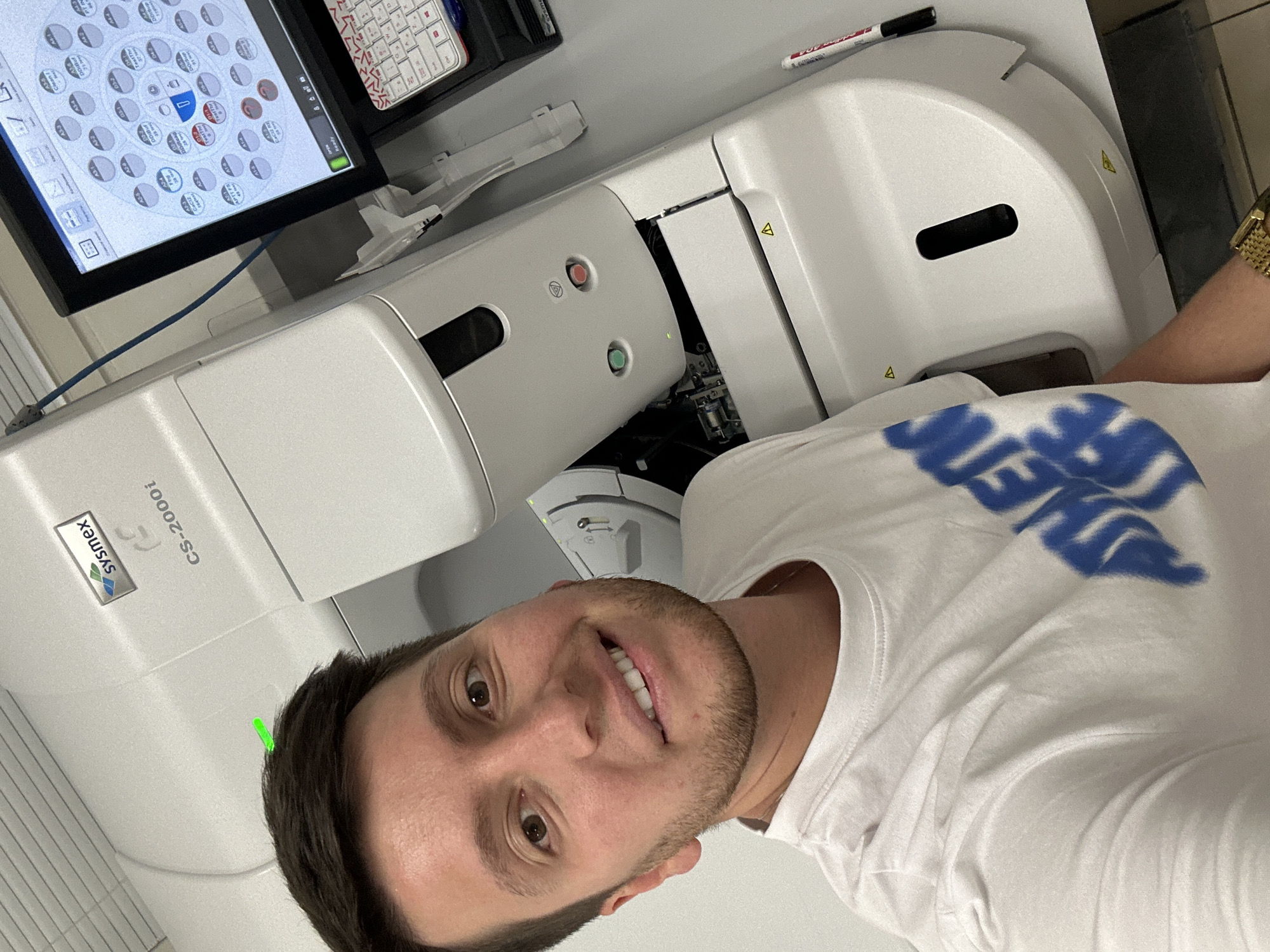 A man with short brown hair and a beard is taking a selfie in a laboratory. He is wearing a white t-shirt with a blue graphic print and a gold watch. Behind him is a large white laboratory machine with the brand name "Sysmex" and model number "CS-2000i". A computer monitor displaying a circular chart with various data points and a keyboard are also visible. A red and black marker is leaning against the machine. The environment appears to be a clean, well-lit laboratory. There is no indication that this image was taken in Moscow, Russia.