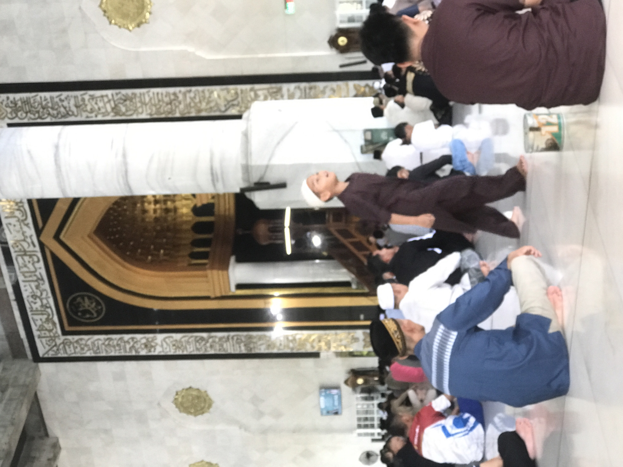 A horizontal, high-angle photograph depicts a group of people praying inside a mosque in Lhokseumawe, Indonesia. The worshipers are barefoot and are kneeling on the floor. A boy in a brown robe is looking upward. The interior of the mosque is ornate, with marble columns and walls featuring Arabic calligraphy and golden geometric patterns.