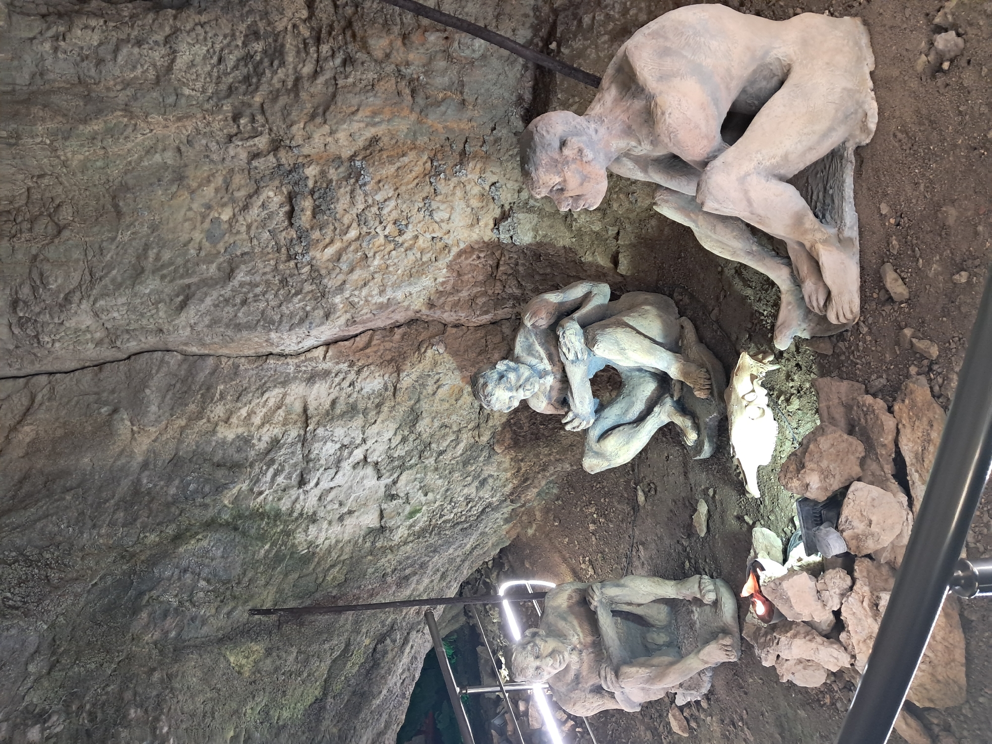 The image shows three human-shaped statues in what appears to be a cave or underground setting. The cave is located in Dryanovo, Bulgaria. The statues are made of a light-colored material, possibly plaster or concrete. One statue is positioned at the top, leaning forward with bent legs. Another statue is in the middle, with limbs intertwined, possibly depicting a struggle. The third statue is at the bottom, also with bent legs. The statues are surrounded by rocky terrain and dirt. A metal rail runs along the right side of the image and a metal beam runs across the top of the image. A light source illuminates the statues and the surrounding area. The walls of the cave are rough and textured.