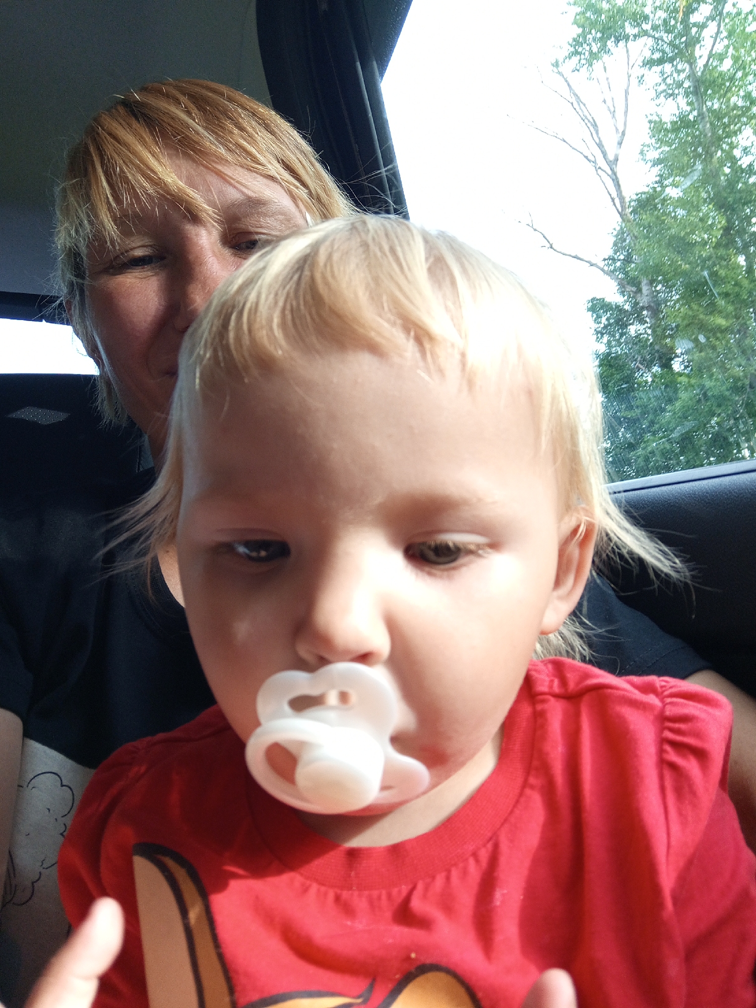 In Buraly, Russia, a close-up, low-angle selfie captures two individuals, an adult and a young child, seated inside a vehicle. The adult, with blonde hair and fair skin, is positioned behind and to the left of the child, with their face partially visible and turned slightly towards the camera. The child, also with blonde hair and fair skin, is in the foreground, their face taking up most of the frame. The child is wearing a red shirt and has a white pacifier in their mouth. The background is blurred, showing foliage and a bright sky, suggesting an outdoor setting visible through a car window. The interior of the vehicle appears to be dark, with hints of black upholstery.