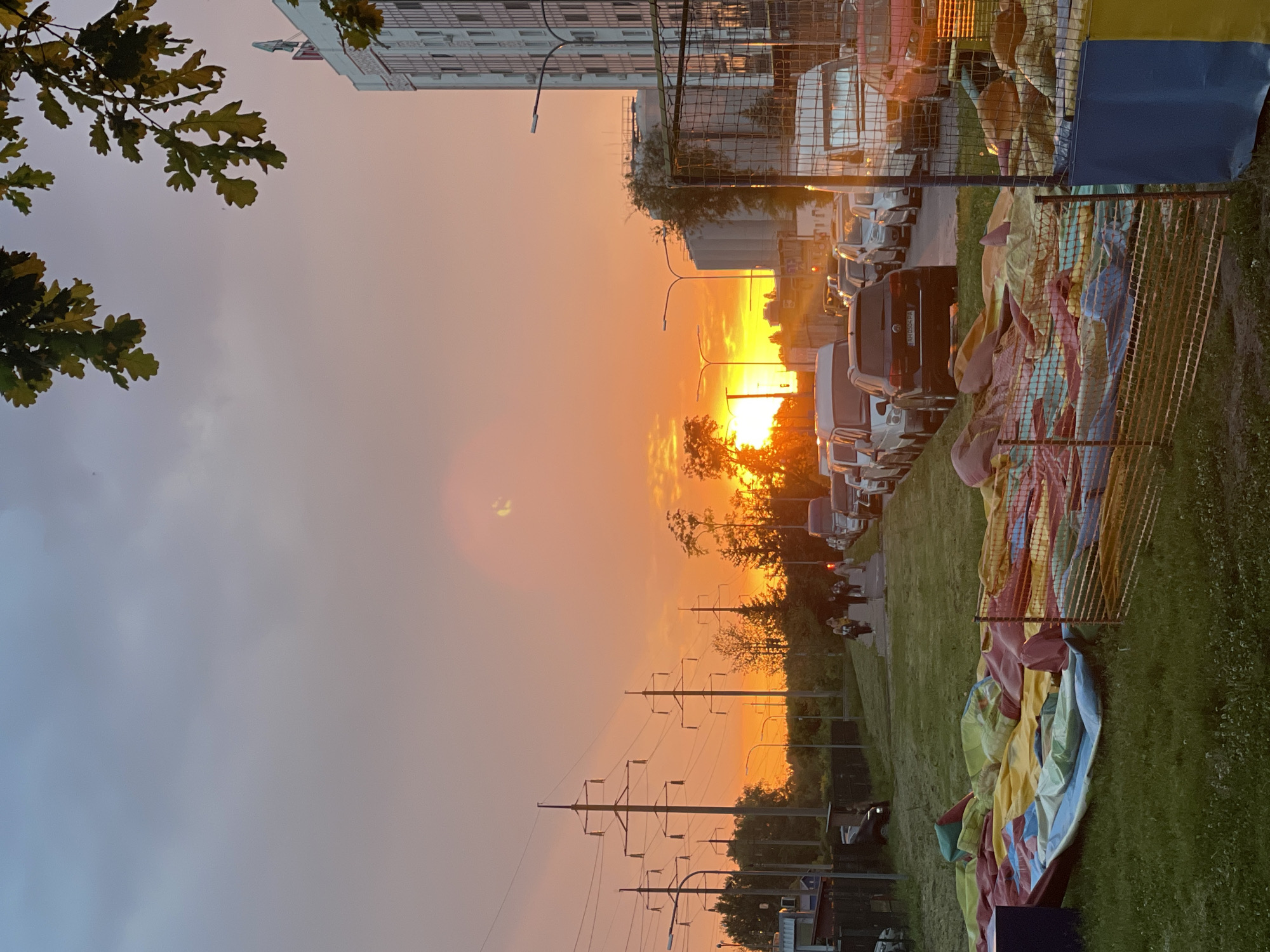 The image captures an urban or suburban street scene at sunset in Rozsoshentsi, Ukraine. The sky is a dominant feature, bathed in brilliant orange and yellow hues as the sun dips below the horizon, casting a warm glow across the landscape. The intense light near the sun gradually fades to a paler orange and then a light blue-grey on the left side, with some scattered clouds visible.
In the upper left corner, dark green oak leaves frame the scene, partially obscuring a tall, light-colored building, likely an apartment complex, which stands against the fading sky. Running across the middle ground are several utility poles with numerous power lines, silhouetted against the bright sunset. Below these, a street is lined with parked cars and vans, their forms largely appearing as dark silhouettes reflecting the golden light. One dark SUV/car in the mid-right shows a visible license plate with the text "AH 0594 PA". A few indistinct figures, possibly people, can be seen in the distance along the street.
On the right side of the image, a strip of green grass borders the road. Lying on this grass, partially covered by a mesh fence, is a large, colorful, deflated or partially deflated inflatable structure. Its sections are visible in shades of yellow, red, blue, and green, suggesting it could be a bouncy castle or similar recreational equipment. Its deflated state implies that an event has either recently concluded or is about to begin. The overall atmosphere is tranquil, characteristic of dusk, with the vibrant sunset contrasting with the shadowed foreground elements.