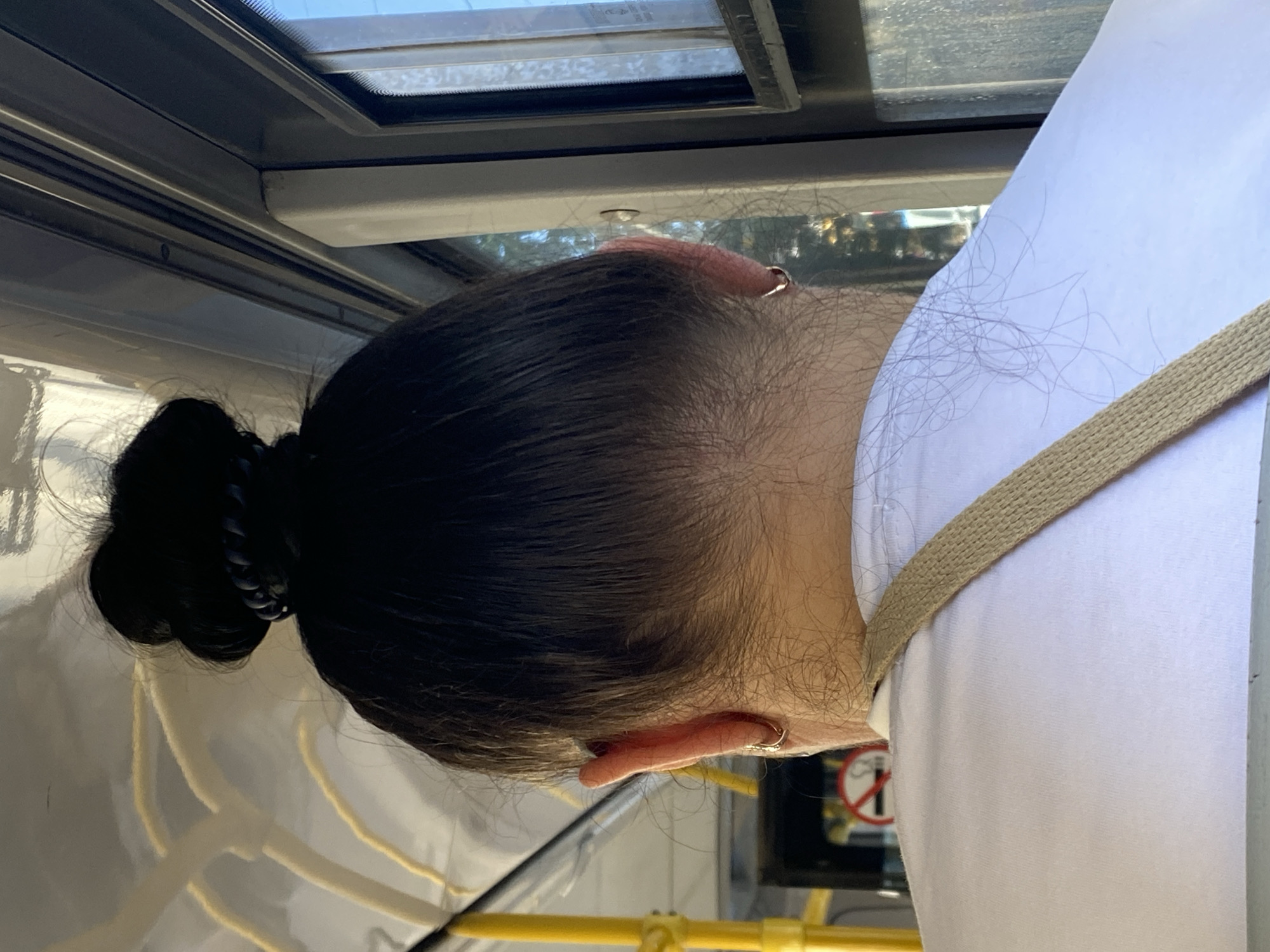 The image shows the back of a person's head and shoulders, viewed from behind, inside what appears to be a public transport vehicle. The person has dark hair styled in a bun on top of their head and is wearing a white t-shirt with a beige strap across the shoulder. The background reveals details of the vehicle's interior, including yellow poles, a window, and grey paneling. The overall lighting suggests an indoor setting during the day. There are no visible elements that specifically indicate the location is Novokuznetsk, Russia.