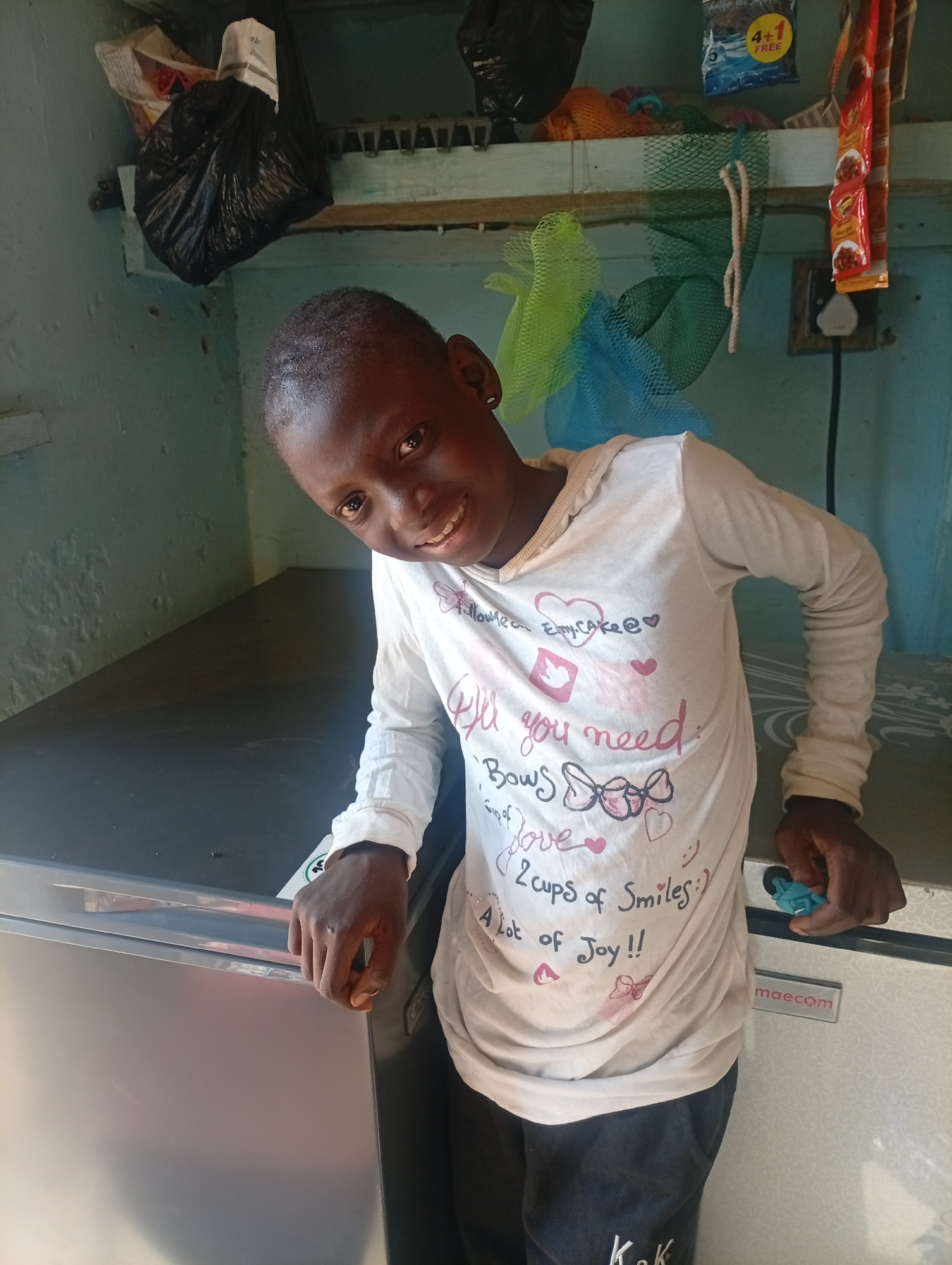 A young, dark-skinned boy is standing in a room. He wears a long-sleeved, white shirt with various motifs and text, including the words "All you need: Bow's love, 2 cups of Smiles, A lot of Joy." He has on dark trousers. The boy is leaning slightly towards the left, resting his hands on a silver-colored, rectangular freezer. Behind him, there is a shelf that holds a black plastic bag, various small items in packaging, and colorful net bags. The wall behind the shelf is a faded turquoise. An electrical outlet with a plug is visible on the right. The scene appears to be indoors, possibly in a small shop.