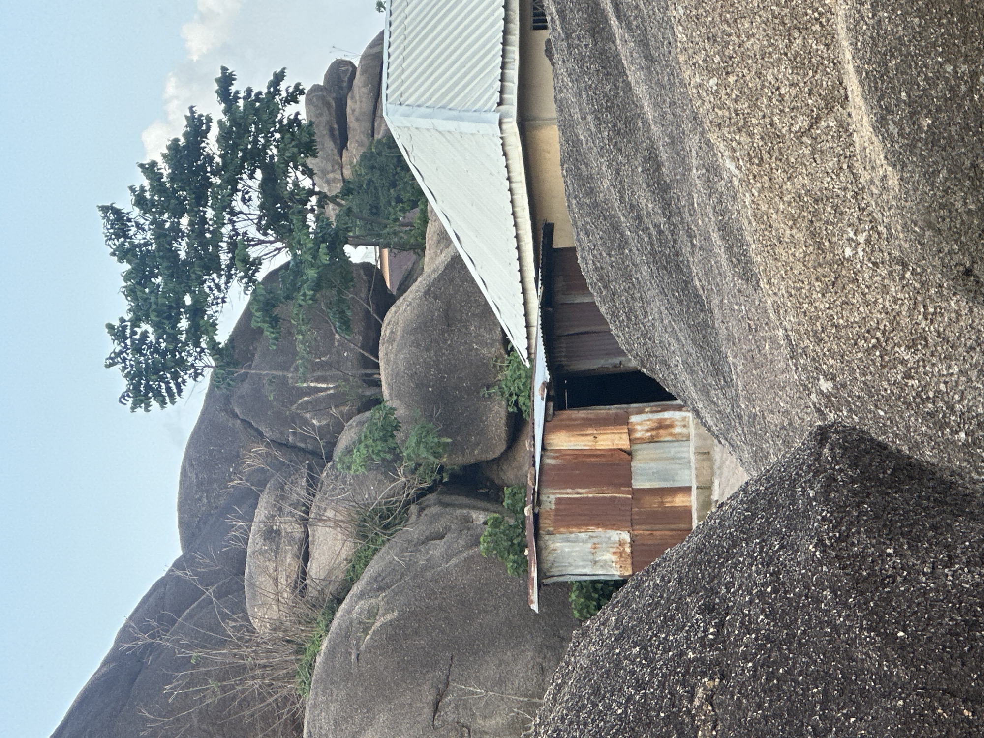 The image depicts a rugged, rocky landscape characteristic of the city of Jos, Nigeria. Large, weathered granite boulders dominate the scene, with sparse vegetation growing in the crevices and on the slopes. In the mid-ground, two structures are visible. The first is a small, makeshift dwelling constructed from corrugated iron sheets, showing signs of rust and wear. Adjacent to it is a more substantial building with a pale yellow exterior wall and a light-colored, corrugated metal roof. The sky above is a pale blue with a few scattered clouds. The foreground is filled with dark, coarse gravelly material, possibly a result of rock quarrying or natural accumulation. The overall impression is one of a natural environment integrated with rudimentary human habitation.