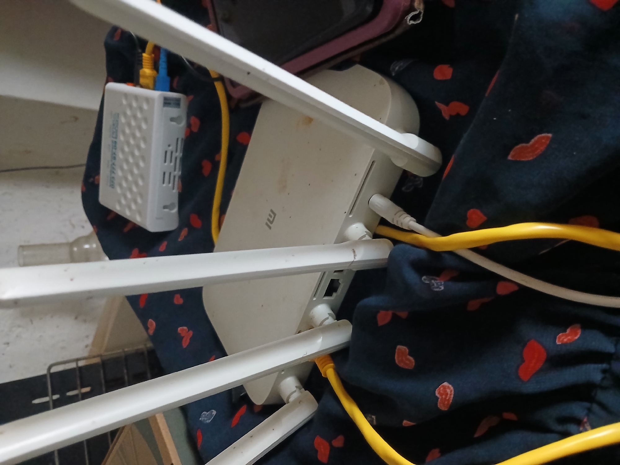 The image shows a close-up of a white Xiaomi router with three antennas, partially obscured by a dark blue fabric patterned with red hearts. Two yellow Ethernet cables are plugged into the router, and a smaller white device, possibly a modem or converter, is visible to the left with blue and yellow cables connected to it. The scene is cluttered, with the router and cables resting on the fabric, which appears to be part of clothing or bedding. In the background, a glass bottle and a metal grate are faintly visible. The lighting suggests it is indoors, with no clear indication of time of day or weather. There is no visible text in the image besides the faint "mi" logo on the router. The overall impression is one of everyday technology set against a soft, domestic backdrop.