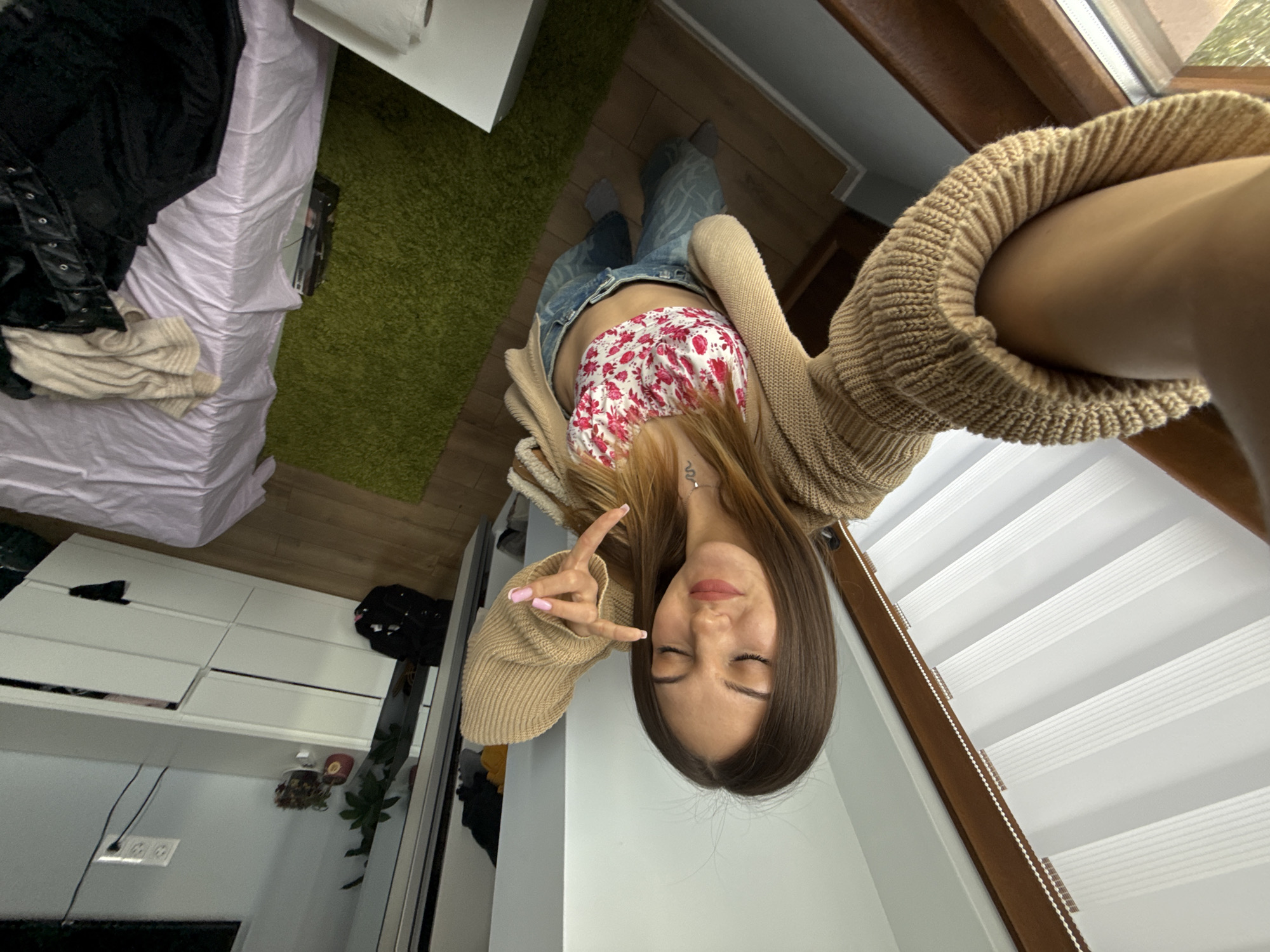 A downward-angled selfie photograph features a young woman with long brown hair wearing a floral top, a tan cardigan, and jeans. She is making a hand gesture. The background includes a room with a bed, a green rug, white drawers, a plant, and a window with white blinds. The location cannot be determined as Sofia, Bulgaria, from the image alone.