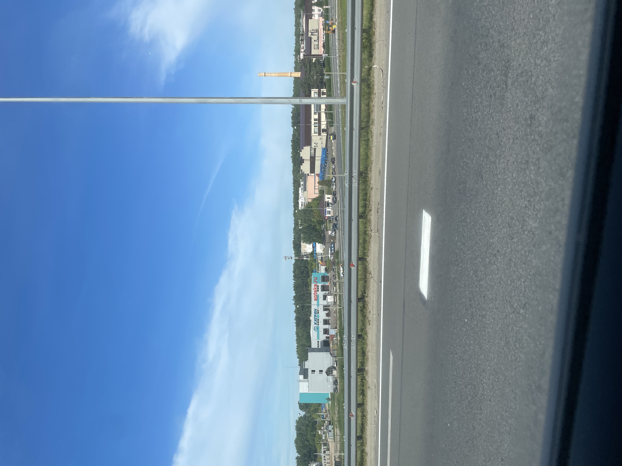 A daytime, eye-level, wide shot captures an industrial area located in Rusanovo, Russia, as viewed from a highway. The foreground features the asphalt road with white lane markings and a bright white rectangular marking. A silver metal guardrail separates the road from a grassy verge. Beyond the guardrail, a collection of buildings stands under a bright blue sky with scattered white clouds. On the left, a modern, light blue and gray building is visible, with other industrial structures and trees in the background. The middle ground shows a long, light-colored building with a blue sign reading "KAPTHEN apteka.ru" and several loading docks. Further to the right, a tall, slender, orange-brown smokestack rises above other industrial buildings. The scene is devoid of people.