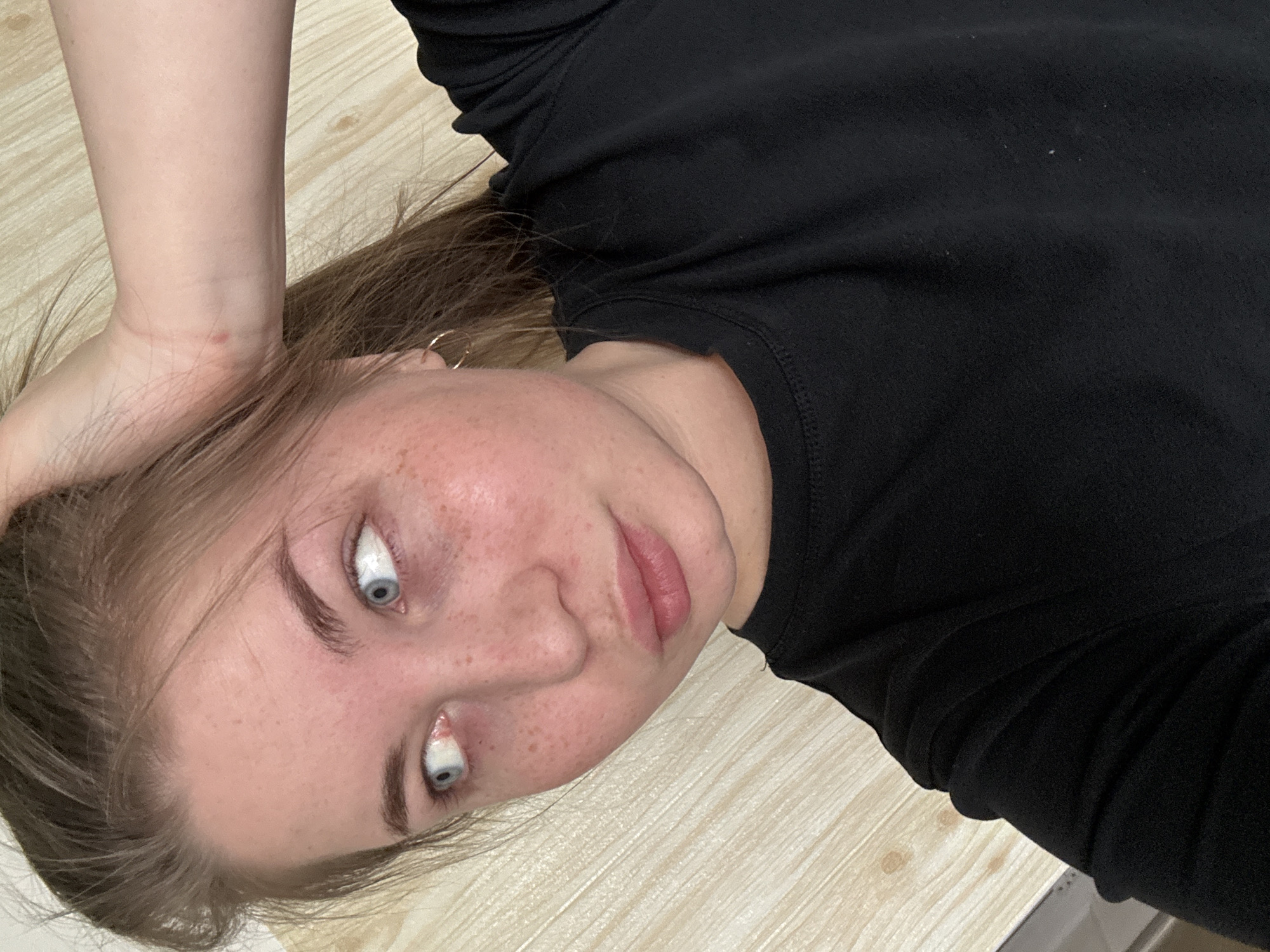 A young woman is captured in a close-up, overhead shot, appearing to be lying down indoors. Her head is slightly tilted, and she looks directly towards the camera. She has light skin with prominent freckles across her nose and cheeks, and light brown or auburn hair that frames her face. Her blue eyes are rolled upwards, revealing a significant amount of white sclera, which gives her expression a striking and somewhat theatrical or playful quality. Her lips are slightly parted.
She is wearing a simple black long-sleeved top, which shows some minor lint or dust particles in closer detail. Her right hand (from the viewer's perspective) is positioned near her head, with a small reddish blemish visible on her wrist. A small hoop earring can be seen on her left ear. The background consists of a uniform, light-colored surface with a distinct wood-grain pattern, likely a wooden floor or a laminate tabletop. The lighting suggests an indoor setting, and no specific time of day or weather can be determined from the image. There is no visible text. The photograph is set in Chaikovsky, Russia.