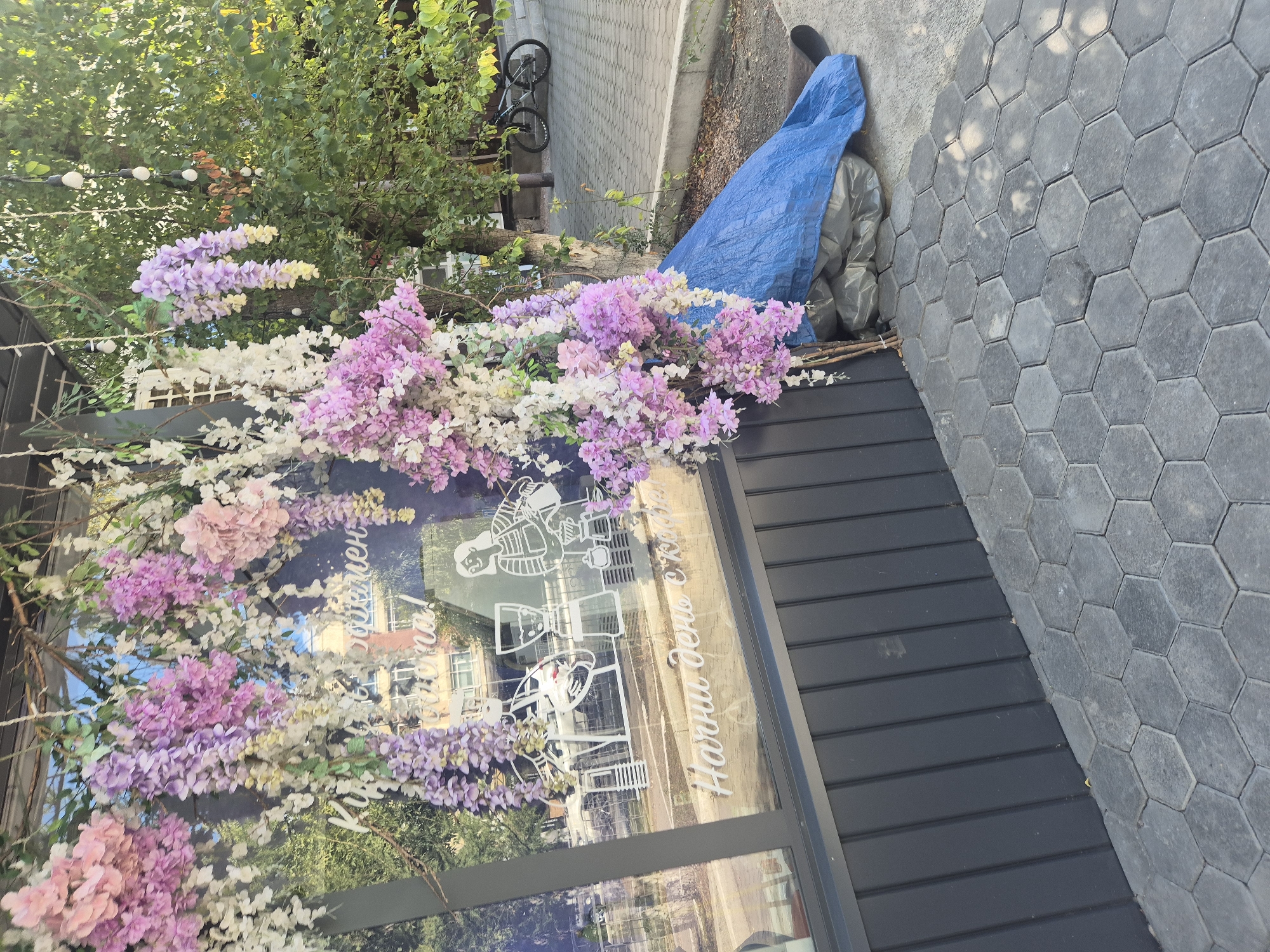 A street-level view captures the exterior of a cafe or shop in Kokshetau, Kazakhstan, decorated with cascading artificial flowers in shades of pink, purple, and white. The flowers hang from an overhead structure, partially obscuring a large window. Etched onto the window are white graphics depicting coffee-making equipment and a person holding a coffee cup. Below this, in white lettering, are the words "Начни день с кофе" (Start your day with coffee).
The building's facade is dark gray with vertical paneling. To the right of the window, a blue tarp covers what appear to be stacked garbage bags or stored items. Beyond the building, a paved area with hexagonal stone tiles leads to a sidewalk and a street. A bicycle is parked near a tree on the right side of the image, with lush green foliage forming a natural backdrop. The scene is illuminated by natural sunlight, casting shadows on the pavement.