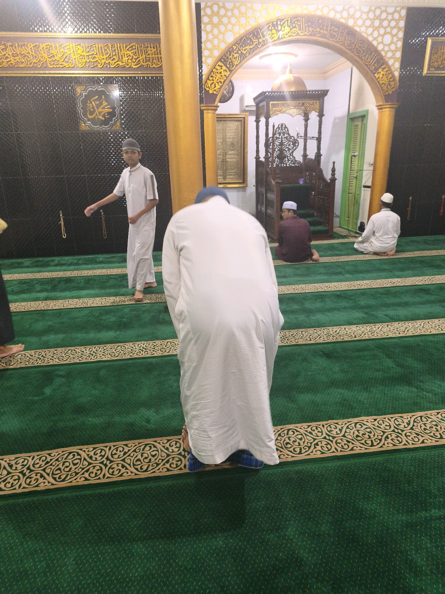 The image shows the interior of a mosque, likely in Samarinda, Indonesia, with green carpeting and decorative elements. In the foreground, a person in a white robe is bowing in prayer, with their back to the camera. To the left, a young boy in a light-colored tunic and cap stands, looking to the side with his arm extended. Further in the background, two men are seated on the carpet in prayer positions. One man wears a maroon tunic and a white cap, while the other wears a white tunic and cap. Behind the seated men, a wooden pulpit or minbar is visible, suggesting a place for religious sermons. The walls are adorned with Islamic calligraphy and intricate patterns. The overall scene depicts a moment of communal prayer within a mosque.