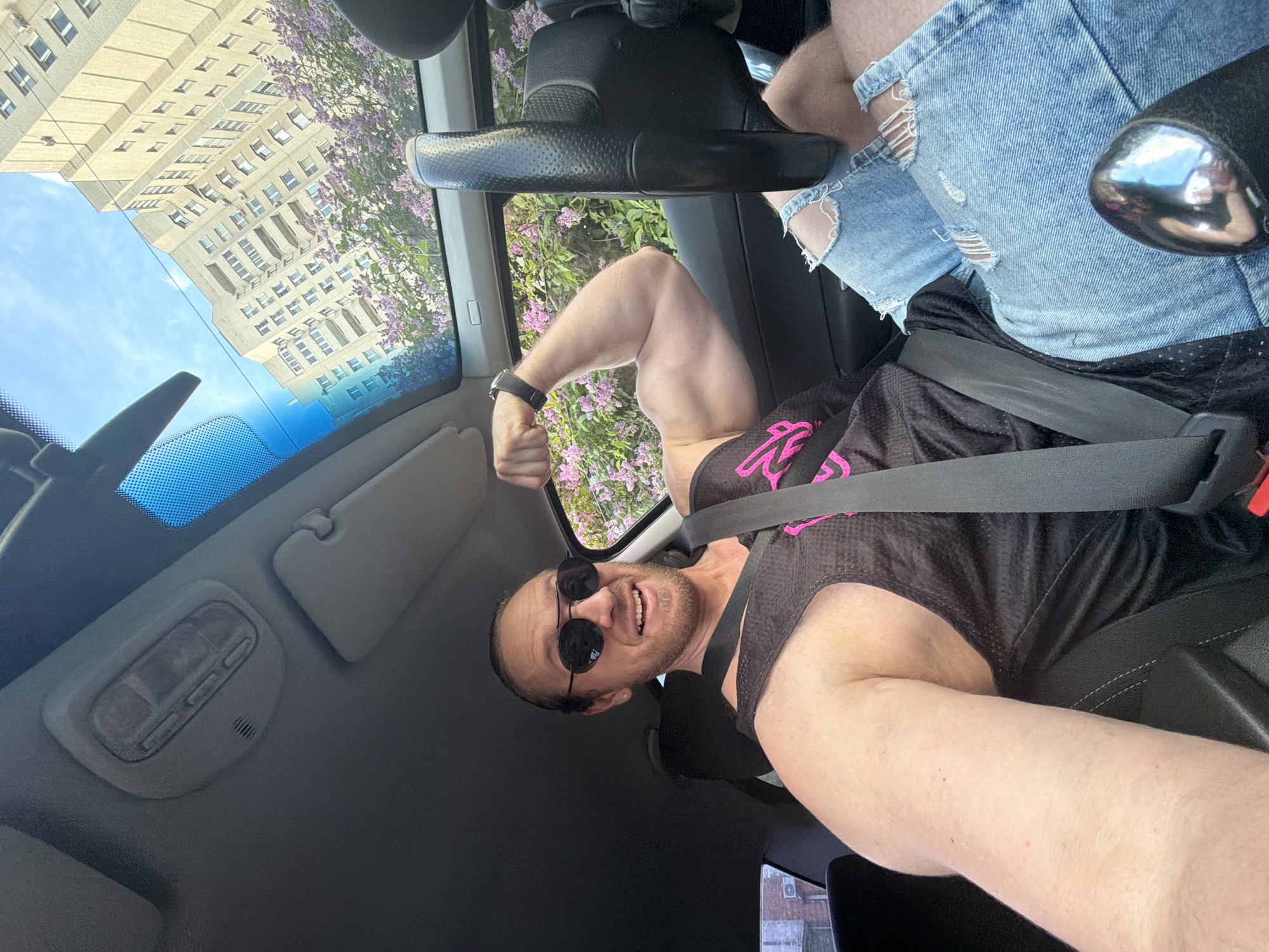 A selfie-style photograph captures an adult male seated in the driver's seat of a vehicle. The man, with light skin, brown hair tied back, a beard, and sunglasses, is smiling broadly with visible teeth while flexing his right bicep. He wears a black athletic tank top with a pink graphic, ripped blue jeans, and a black wristwatch on his left wrist. A seatbelt is fastened across his chest.
The car's interior is grey, featuring an overhead light console with buttons and a small grille, a folded sun visor, and a panoramic sunroof. Through the sunroof and windows, a clear blue sky with some clouds is visible, along with the upper portion of a tall, light-colored residential building characterized by numerous windows and balconies, consistent with post-Stalinist architecture found in Saint Petersburg, Russia. Blooming lilac bushes with purple flowers are visible outside the car, indicating spring or early summer. A black steering wheel and a metallic gear shift knob reflecting light are partially visible in the lower right of the frame.