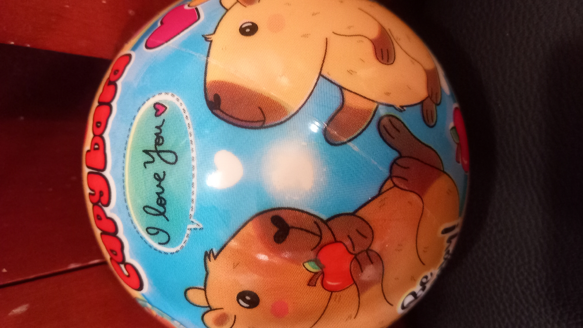 The image shows a round object, likely a toy ball, decorated with cartoon capybaras and the text "Capybaras" and "I love You". One capybara is holding a red apple. The ball has a blue background with white hearts. The object is placed on a dark, textured surface, possibly a piece of furniture. The background is blurred and appears to be a reddish-brown surface. There is no indication of the city of Kos, Greece in the image.