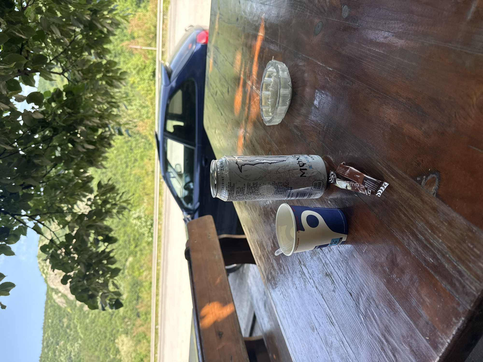 The image depicts a scene on a wooden picnic table. Visible on the table are a Monster energy drink can (white), a disposable cup with a blue design, two small chocolate bar wrappers, and a clear glass ashtray. The table appears to be outdoors. In the background, a blue car is parked beside a road. The location features greenery, including trees and a hillside, consistent with a landscape near Paidushko, Bulgaria. No people are directly visible in the frame.