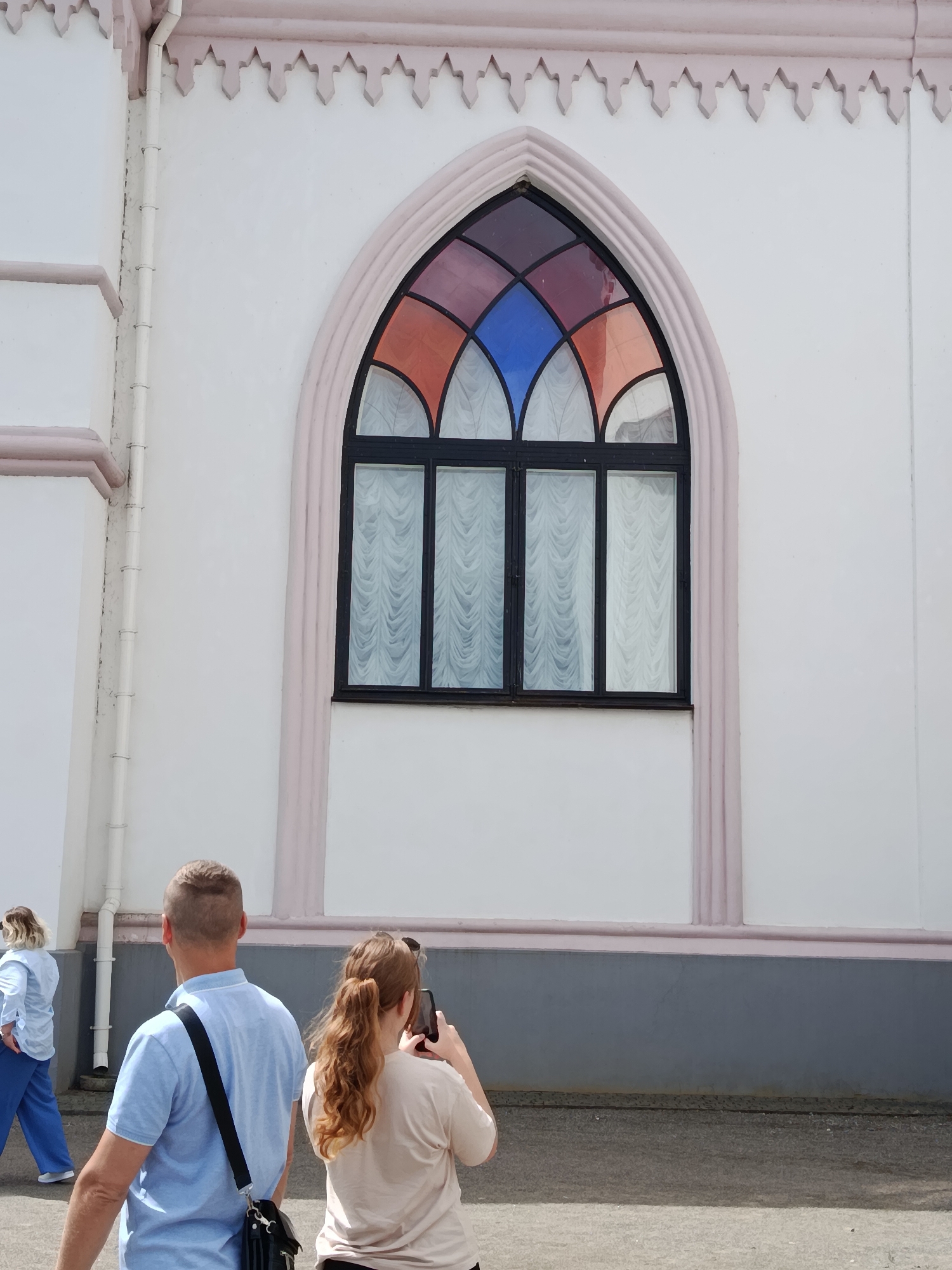A group of people are seen walking in front of a white building with a decorative pink trim and a stained-glass arched window. The building is located in the city of Квасевіцкі сельскі Савет, Belarus. In the foreground, a man with a short haircut, wearing a light blue polo shirt, walks away from the camera with a black bag slung over his shoulder. To his right, a woman with long, wavy red hair, wearing a light-colored t-shirt, holds a black smartphone to take a picture. Further to the left and in the background, a woman with blonde hair, wearing a light blue shirt and blue pants, walks away from the camera. The ground is paved with asphalt.