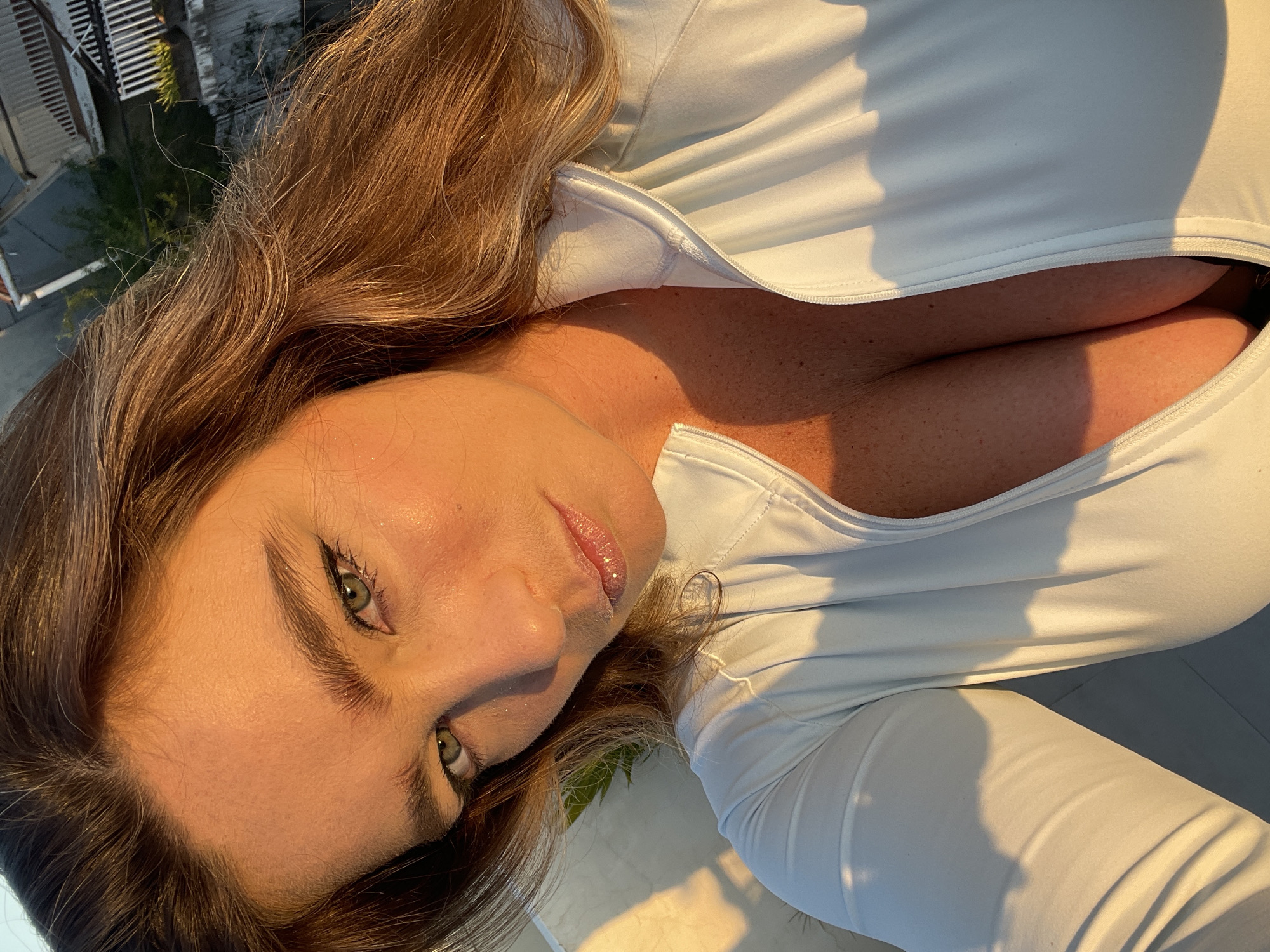 A woman with long, wavy brown hair is captured in a close-up, head-and-shoulders shot, likely taken outdoors in North Kuta, Indonesia, as indicated by the visible rooftop in the background. She is wearing a white, zippered athletic top that is partially unzipped, revealing her cleavage. The sunlight highlights her facial features, particularly her green eyes and sparkling lips. Her skin appears to have a warm glow from the golden hour light. The background shows a residential rooftop with some greenery, characteristic of a tropical urban environment. The overall impression is a candid, sun-drenched selfie.