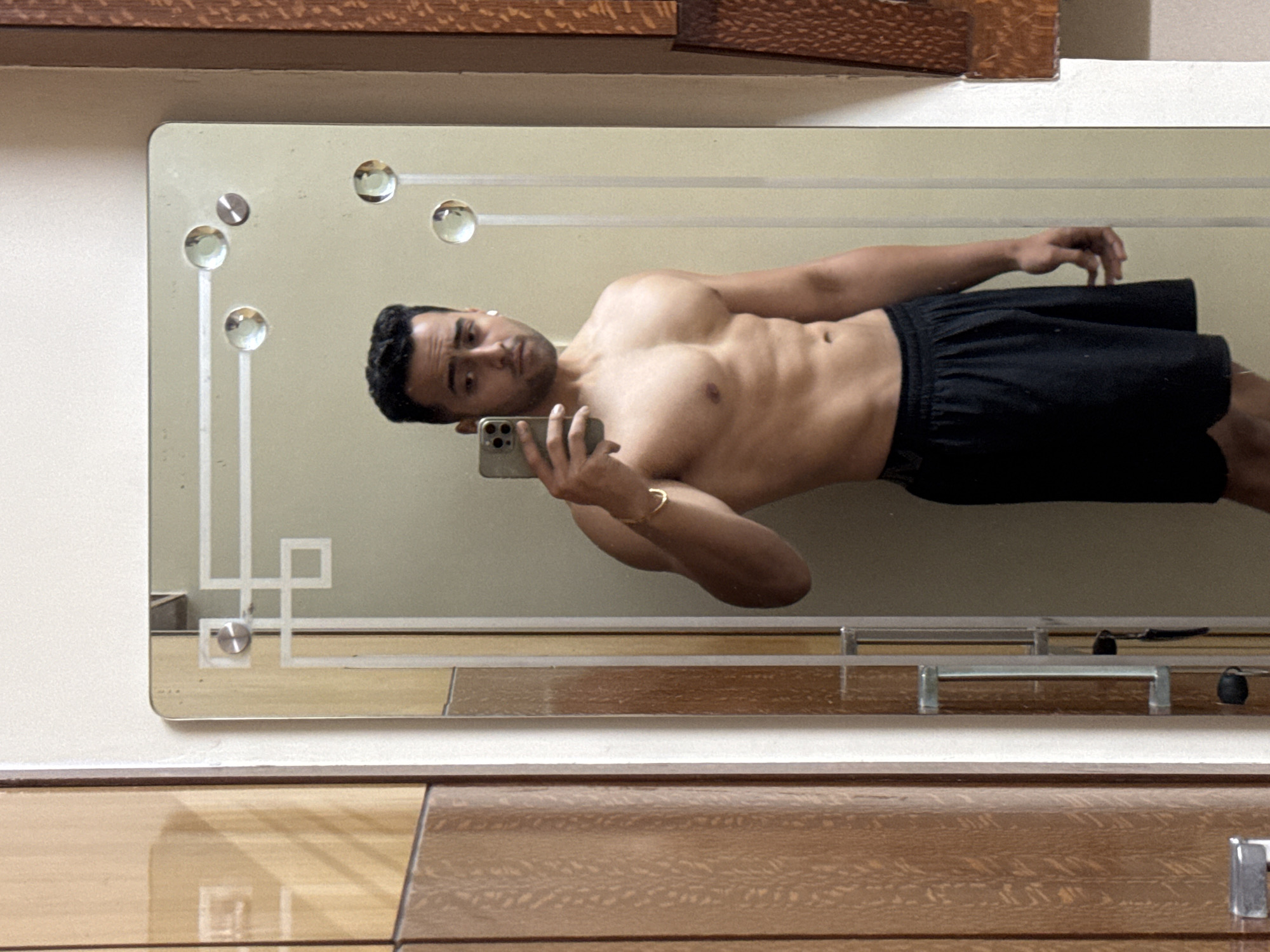 A man is taking a mirror selfie. He is shirtless, wearing black shorts, and holding a phone in his left hand. He appears to be in a bathroom based on the mirror's reflection of a vanity or cabinet. The location in Kolkata, India, cannot be determined based on the provided image.