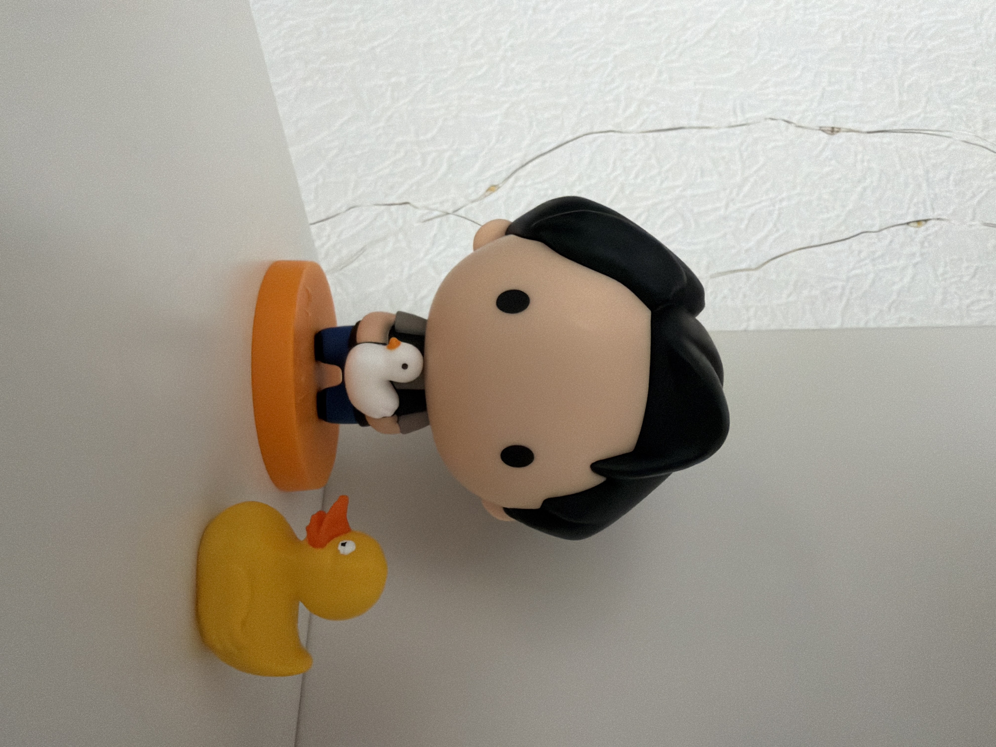 A figurine of a person with black hair and large eyes, wearing a gray shirt and blue pants, stands on an orange circular base. The figurine is holding a small white duck with an orange beak. To the right of the figurine is a yellow rubber duck with an orange beak and white eyes. The objects are placed on a white surface, with a textured white wall behind them. Thin, clear strings with small lights are visible on the wall. The scene is indoors, with soft lighting and a shallow depth of field, suggesting a shelf or desk in an apartment in Kyiv, Ukraine.