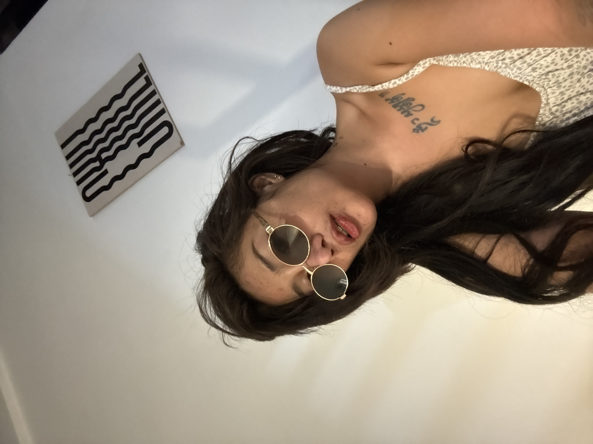 An overhead shot captures a person lying down indoors on a light-colored surface, likely a floor or bed. The subject is a young woman with long dark hair, wearing round gold-rimmed sunglasses and a light-colored, possibly white or cream, patterned top with thin straps. Her head is angled, with her gaze directed towards the camera, and her lips are slightly parted, revealing a hint of her tongue.
A prominent tattoo in a foreign script, resembling Sanskrit or a similar language, is visible on her upper right chest, extending towards her shoulder. Her skin shows some minor blemishes. To the left of her head, a book or magazine with a light-colored cover rests on the same surface. The cover features stylized black text in a wavy, block-like font, which appears to spell "BUILD" or a similar word, although its abstract nature makes definitive identification challenging. The overall lighting is bright and even. The image has a casual, possibly playful or stylish, aesthetic.
