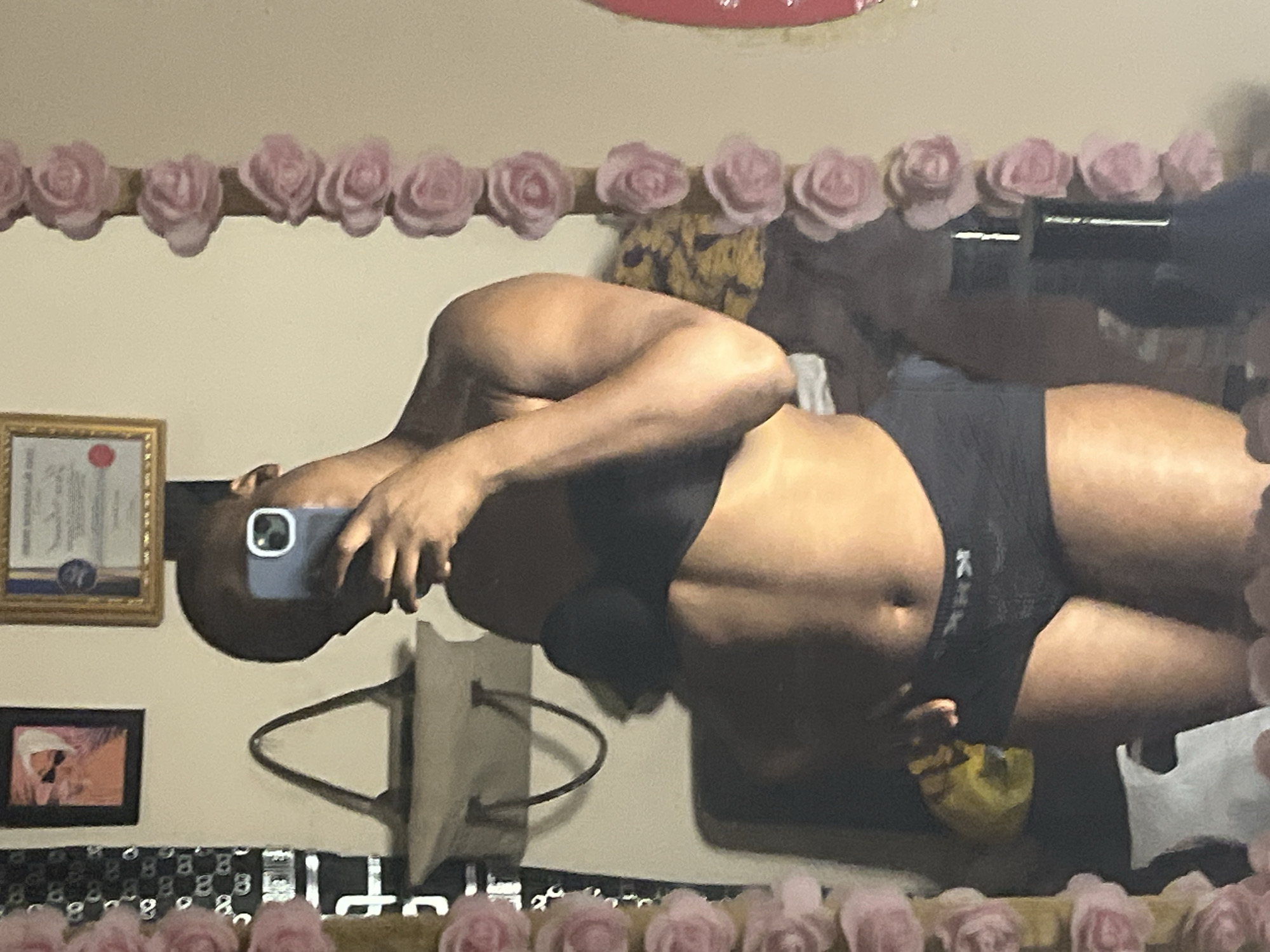 A person in a black bra and black shorts is taking a selfie in front of a mirror. The mirror is decorated with pink flowers around the edges. Behind the person, the mirror reflects a wall with a framed certificate, a small framed picture, and a bag hanging on the wall. The bottom of the mirror's reflection shows a black and white patterned surface. There is no information that indicates the city of Jalingo, Nigeria.