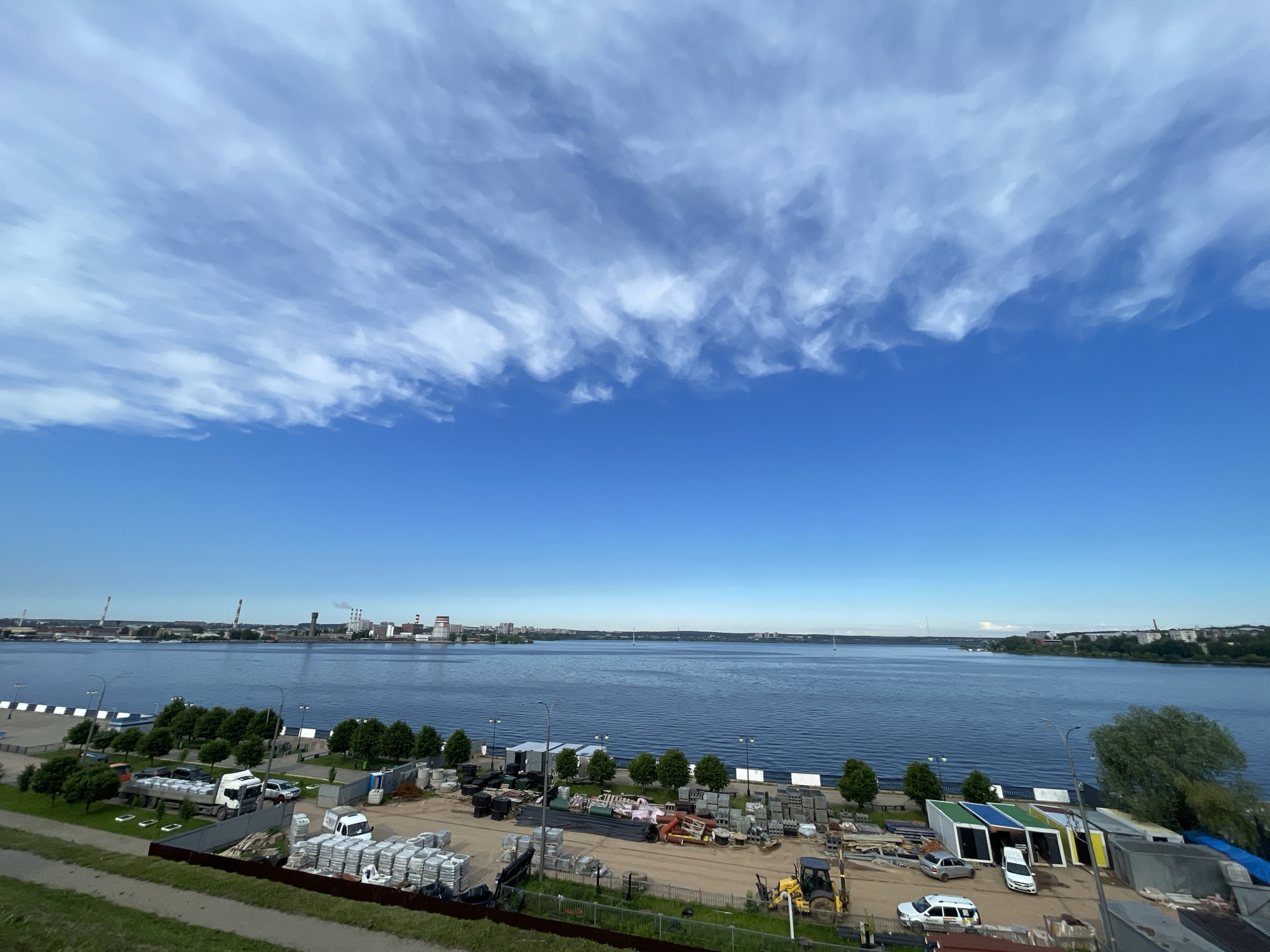 The image captures a wide panoramic view of a calm body of water under a vast blue sky. The upper half of the sky is adorned with a sweeping band of wispy, stretched white clouds, suggesting movement or high winds at altitude. Below this, the sky transitions to a clear, brilliant blue towards the horizon.
In the foreground, a grassy embankment slopes down to a fenced-off industrial or construction storage yard. This yard is densely packed with various materials, including stacks of bagged goods (possibly cement or aggregates), concrete blocks, pipes, and metal components. Several vehicles are present: a large white semi-truck loaded with bagged materials, a yellow backhoe or excavator, a white van, and a silver car. Alongside the water's edge, a row of small trees lines a paved walkway or embankment. Further right in the foreground, several colorful, modular container-like structures (blue, green, yellow, white) are visible.
The midground is dominated by the wide expanse of dark blue water, which shows only minor ripples. On the opposite bank, distant city infrastructure is visible. To the left, a cluster of industrial buildings with multiple smokestacks, some emitting plumes, indicates a power plant or factory complex. Towards the right, the silhouette of a long, low bridge can be seen spanning the water in the far distance, connecting distant landmasses. The overall scene suggests a sunny day with bright, even lighting, likely in the morning or early afternoon. No explicit human activity or visible text is discernible.