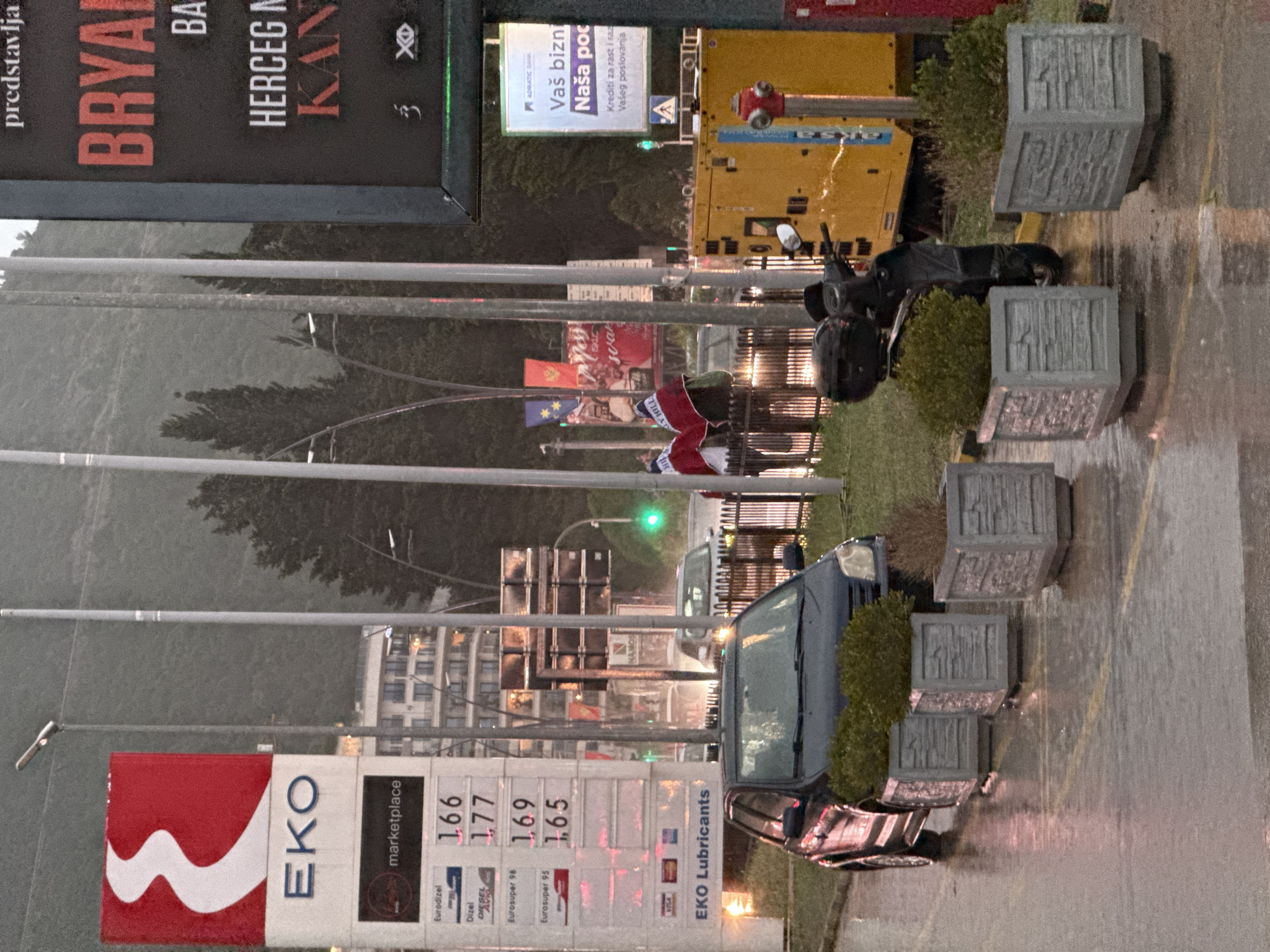 The image, captured at dusk or night in Budva, Montenegro, presents a wet, urban street scene, likely after rain, with reflections visible on the asphalt.
In the foreground, a prominent "EKO" gas station sign displays fuel prices: Eurodizel 1.66, Dizel 1.77, Eurosuper 98 1.69, and Eurosuper 95 1.65. The sign also features "marketplace" and "EKO Lubricants" branding. A dark-colored vehicle, possibly a sedan or van, is parked on the wet ground, flanked by several concrete planters containing small bushes. Further into the scene, a black scooter is visible, along with additional planters and greenery.
Towards the mid-ground, a person dressed in a distinctive red and white outfit, suggestive of a Santa Claus costume or a festive uniform, stands near a pole adorned with multiple flags. These include the flag of Montenegro, the European Union flag, and an unusual flag bearing the text "REPUBLIC OF KLY HILL."
The background is dominated by large advertising billboards. One billboard promotes "ADRIATIC BANK" with text in Montenegrin/Serbian/Croatian that translates to "Your business," "Our help," and "Loans for growth and development of your business." Another large, dark billboard at the top left advertises "BRYAN" and "HERCEG N," with smaller text "predstavlja" (presents) and "KAN," likely indicating a movie or event promotion. A piece of bright yellow industrial equipment can also be seen further down the street. Traffic lights and streetlights provide illumination, contributing to the atmospheric reflections on the wet road surface.