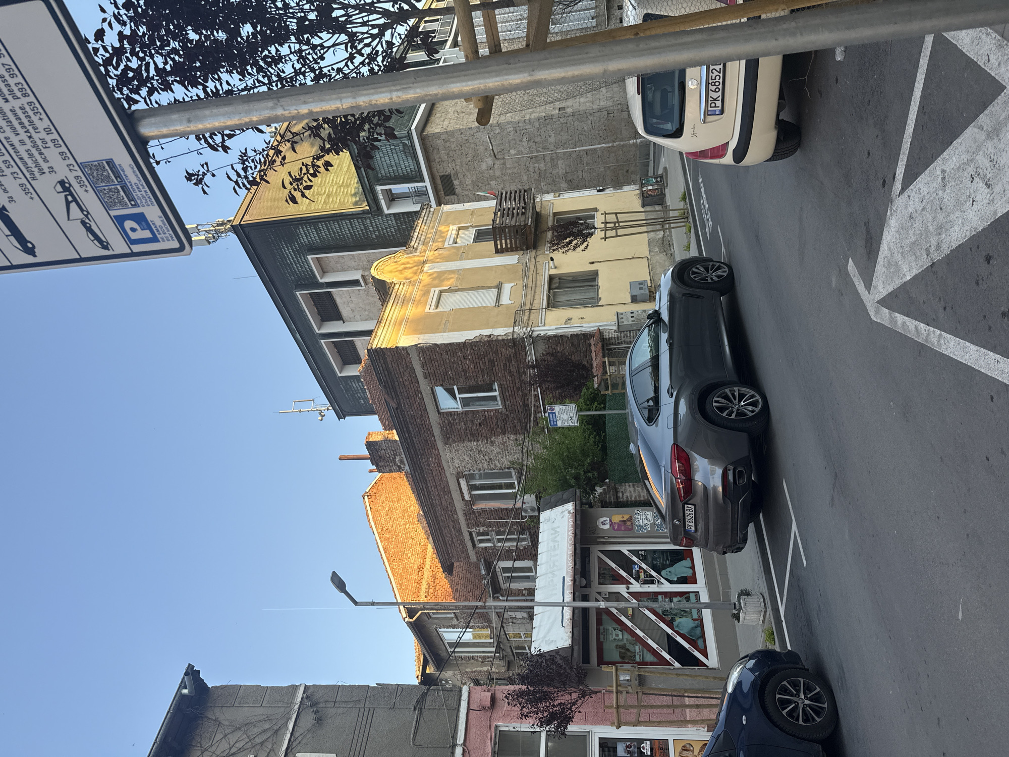 The scene depicts a street in Blagoevgrad, Bulgaria, with parked cars and buildings on either side. A gray BMW X6 (license plate PK 8626 BX) is parked along the street. A white Fiat (license plate PK 6852) is seen in front of the BMW. A dark blue car is also parked.
A parking sign with the text "+359 73 59" is partially visible. The buildings are a mix of architectural styles, with brick, stucco, and tile roofs. A shop called "ПРИЯТЕЛИ И АКСЕСОАРИ" ("Friends and Accessories") is along the street. A street lamp is visible.