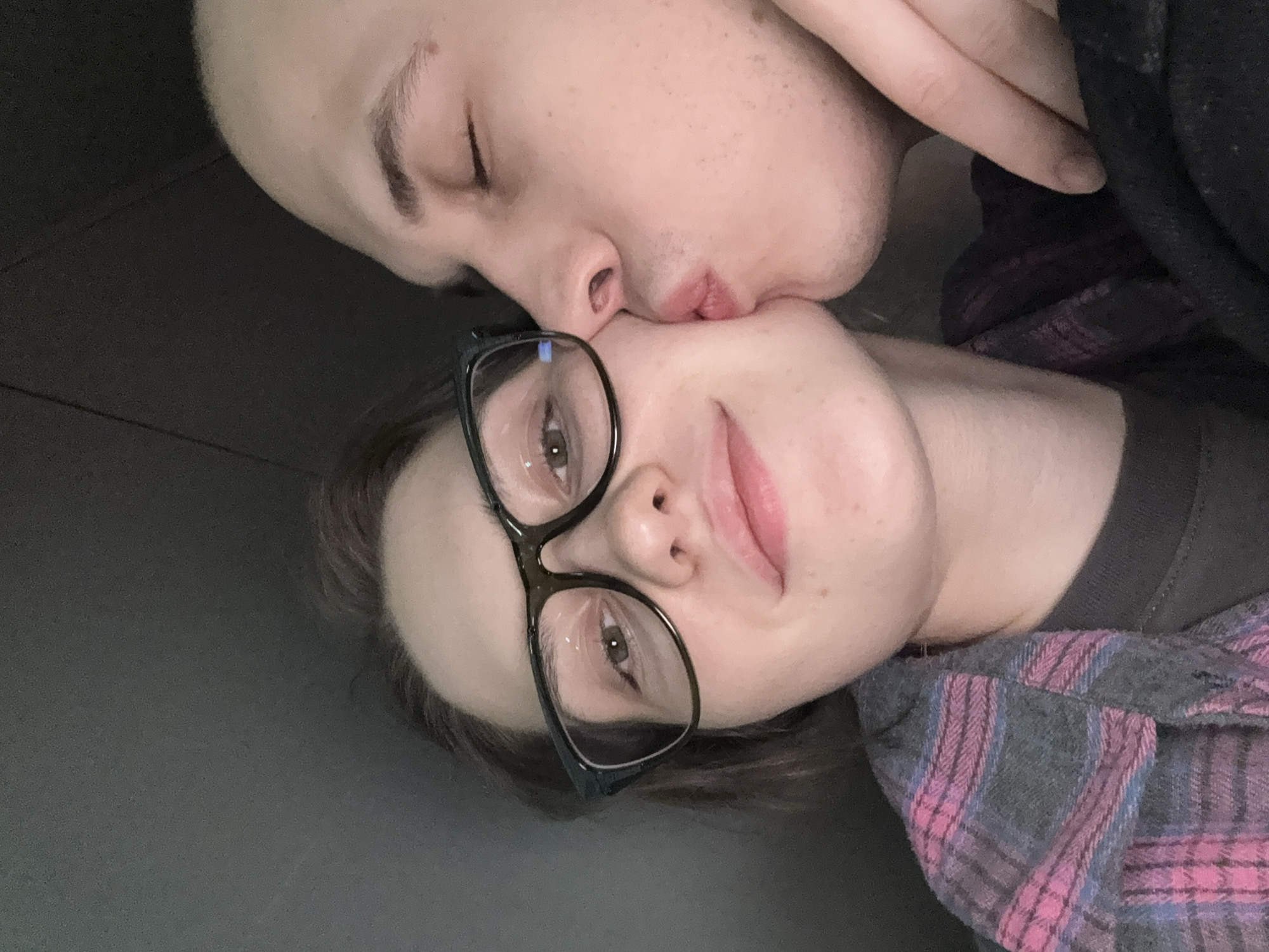 This is a close-up, low-angle selfie of a young couple. The woman on the left is wearing glasses and a pink and gray plaid shirt. She is looking directly at the camera with a slight smile. The man on the right is kissing her cheek. The background is dark and out of focus. The image appears to be taken indoors, possibly in a car. There is no visible indication of the location being in Moscow, Russia.
