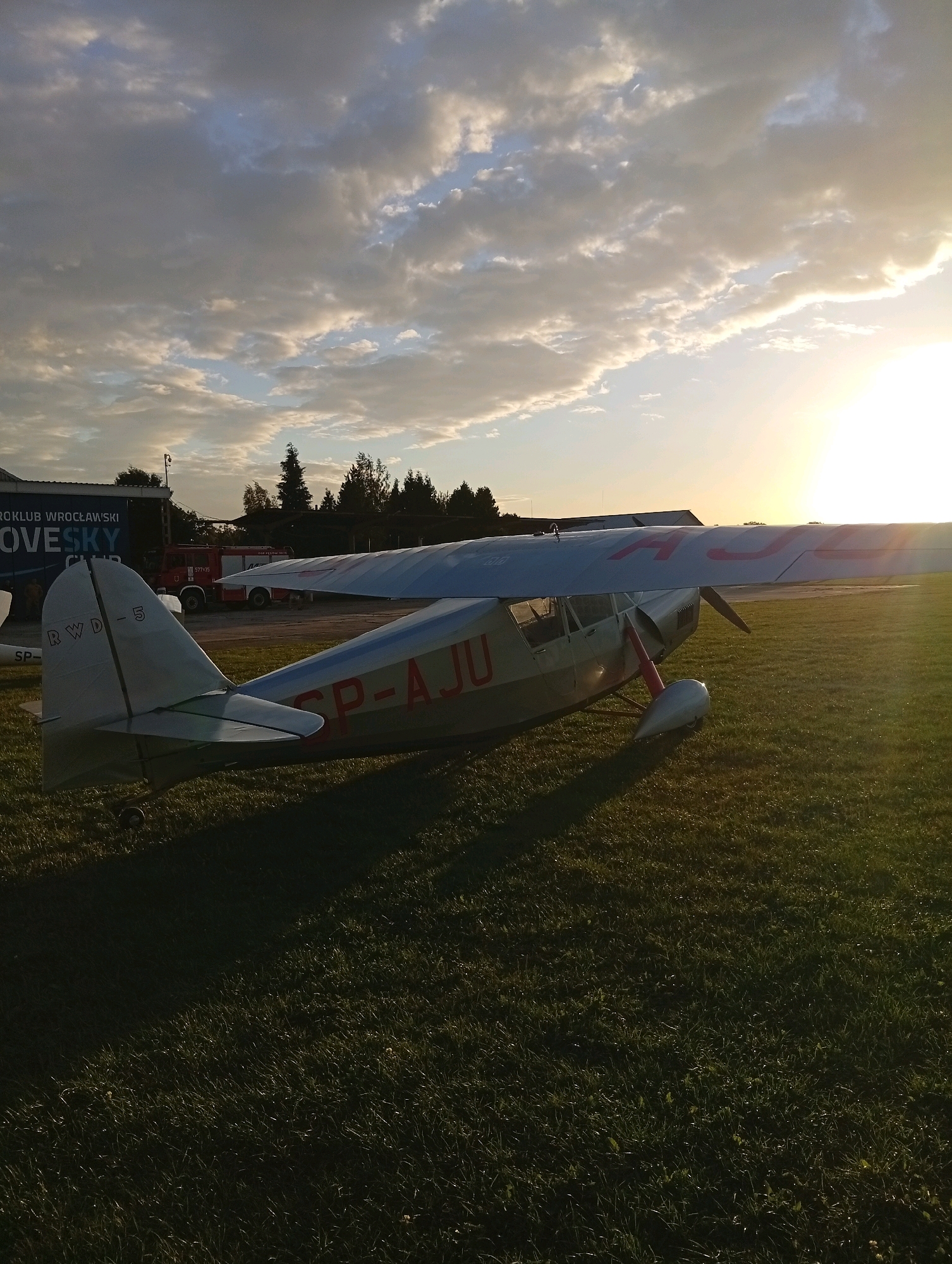An aerial photograph shows a small, light-colored airplane with red markings parked on a grassy field. The airplane has the registration "RWD - 5" on its tail and "SP-AJU" on its wing. The image is taken at sunset, with the sun's rays illuminating the sky and casting long shadows across the field. In the background, a building with text that appears to be "WRÓCKI WROCLASKI" and "OVESKY CLUB" is visible, along with a red fire truck. Trees line the horizon, and a bright sky with scattered clouds completes the scene. The location is the Szewce, Poland aerodrome.