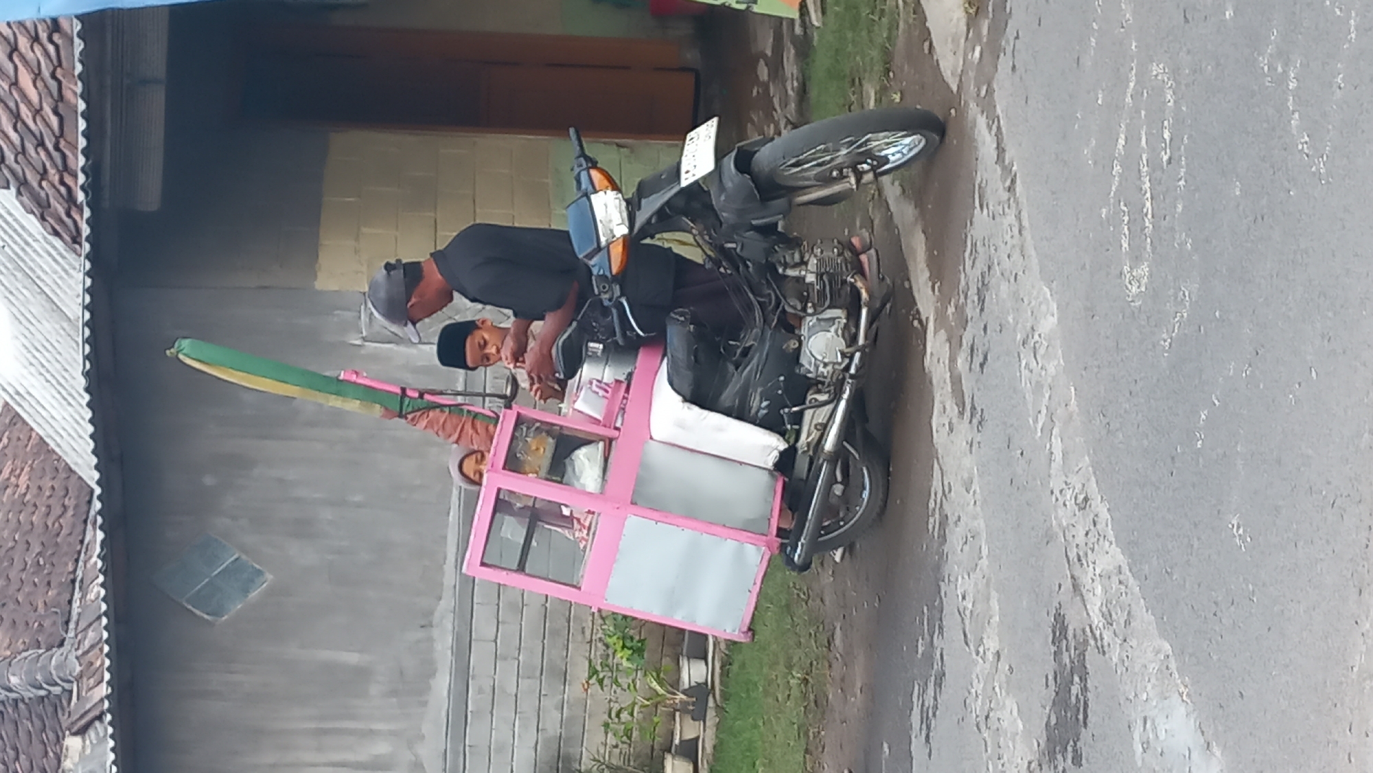 In the city of Pare, Indonesia, a motorcycle with a pink food cart attachment is parked on a paved road. The food cart is designed to be pulled by the motorcycle and has glass display cases. Inside the cart, two children are visible. One child, a boy wearing a black cap, is interacting with an adult male standing next to the cart. The adult male is also wearing a black cap and a dark shirt. The other child, a girl wearing a headscarf, is seated inside the cart. The background shows a building with a tiled roof and a grey wall. The scene depicts a commercial activity, likely street food vending, with a family or associates involved.