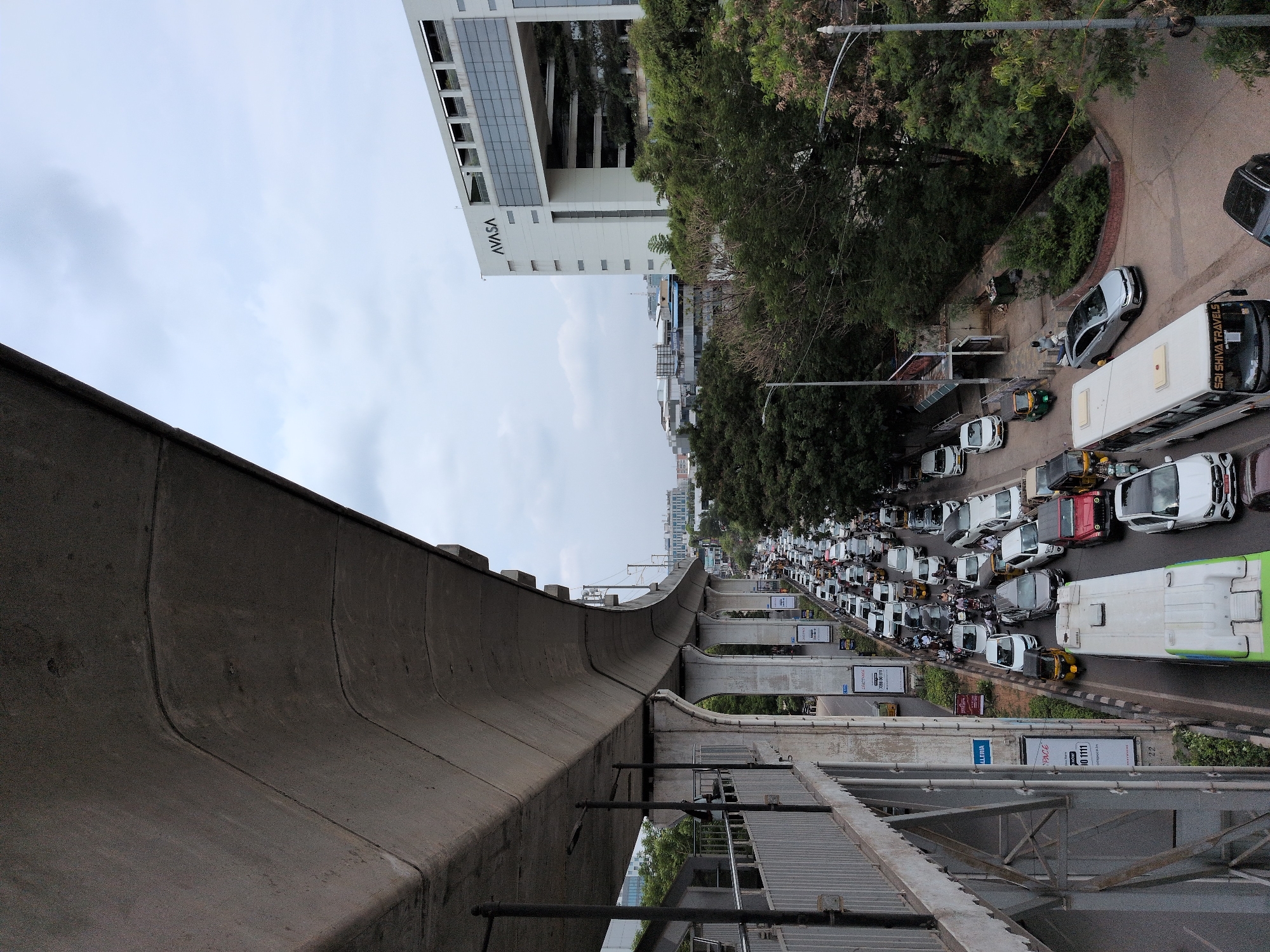 The image shows a heavy traffic jam on a multi-lane road in Hyderabad, India, under an elevated highway or metro line. The road is filled with cars, motorcycles, auto-rickshaws, and a bus. The vehicles are bumper-to-bumper, indicating a standstill situation. On the right side of the road, there is a modern, white building with the prominent "AVASA" logo. Trees and greenery are visible on the sides of the road, and the sky is overcast. The elevated structure on the left is made of concrete and appears to be a pedestrian walkway or part of the metro infrastructure, with a metal railing along its edge. Advertisements are posted on vertical poles along the road. The scene depicts a typical urban scenario of traffic congestion in a bustling Indian city.