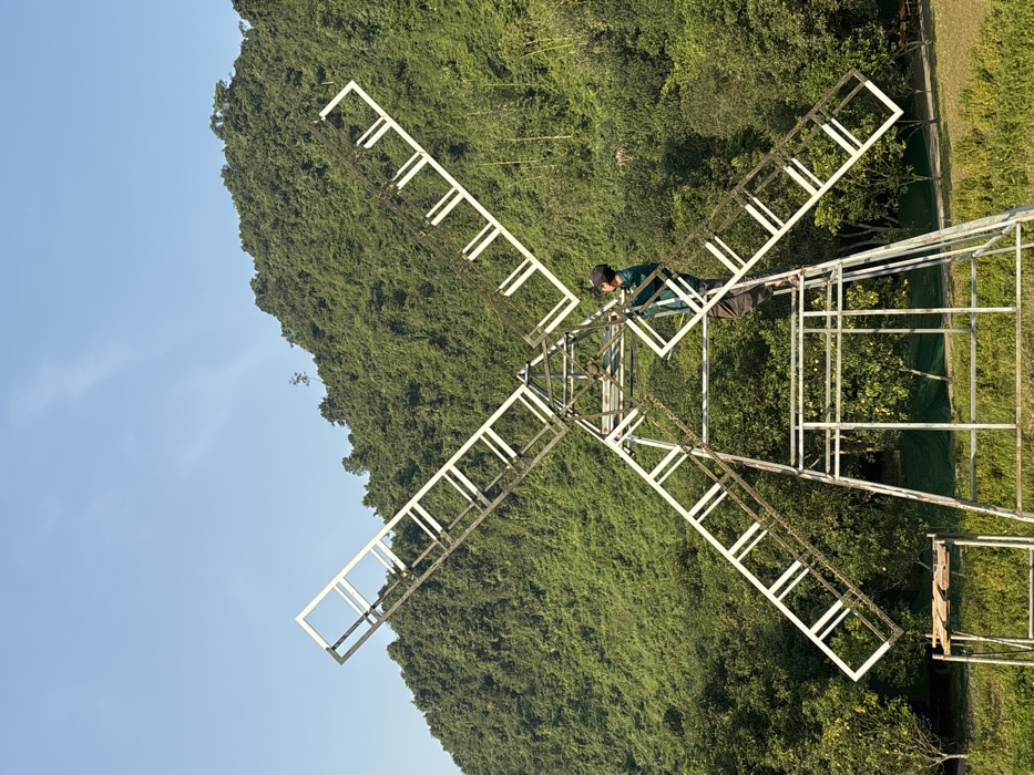A solitary male figure is positioned on a large, skeletal, metal windmill-like structure in a natural outdoor setting within Yên Sơn Ward, Vietnam. The structure, appearing to be constructed from white or light-colored rectangular metal tubing, features four prominent blades arranged in an 'X' configuration, with darker internal bracing. The individual, wearing a dark cap, a teal long-sleeved shirt, and dark pants, stands barefoot on a horizontal beam near the lower center of the structure, facing a blade section and holding onto it with both hands, suggesting active work or inspection. The immediate surroundings include low green vegetation and trees, with a small, simple wooden table or stand visible to the left of the structure's base. In the background, a large, steep mountain or hill heavily covered in dense green foliage dominates the upper half of the image, beneath a clear, light blue sky.