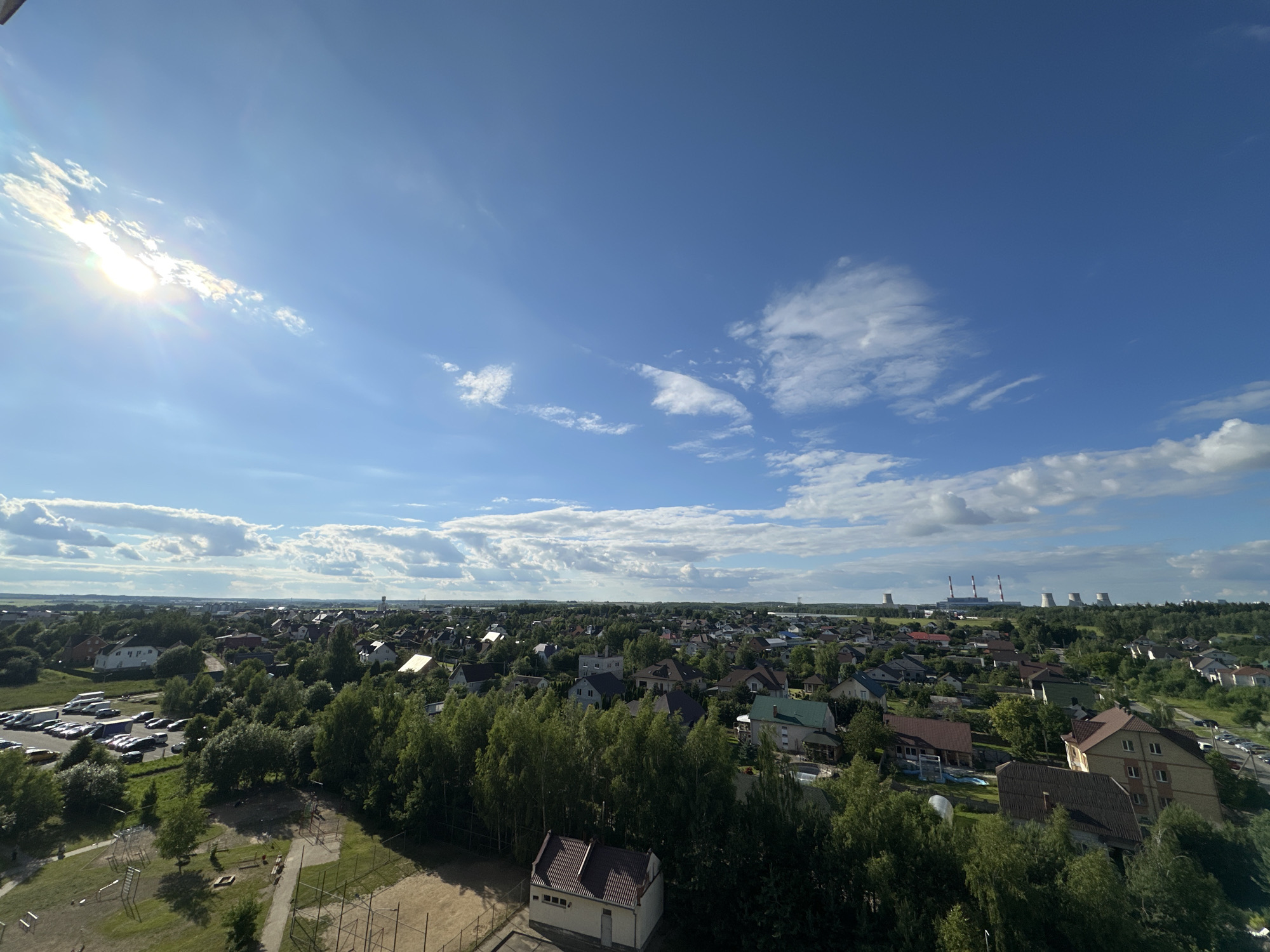 The image is an aerial view of a suburban area, likely in the vicinity of Шчомысліцкі сельскі Савет, Belarus, under a bright blue sky with scattered white clouds and the sun shining. The foreground shows a grassy area with a children's playground, including climbing structures and sandboxes, bordered by a chain-link fence. To the left, a parking lot is filled with numerous cars. Beyond this, a densely wooded area with various trees stretches across the mid-ground. Scattered among the trees are numerous residential houses, most with peaked roofs, and some featuring visible pools. In the background, a vast expanse of similar residential buildings and trees continues to the horizon. Further in the distance, several industrial structures, including cooling towers and smokestacks, are visible, suggesting a power plant or industrial complex. The overall scene depicts a developed residential area with a mix of suburban housing and natural greenery, under a clear, sunny sky.