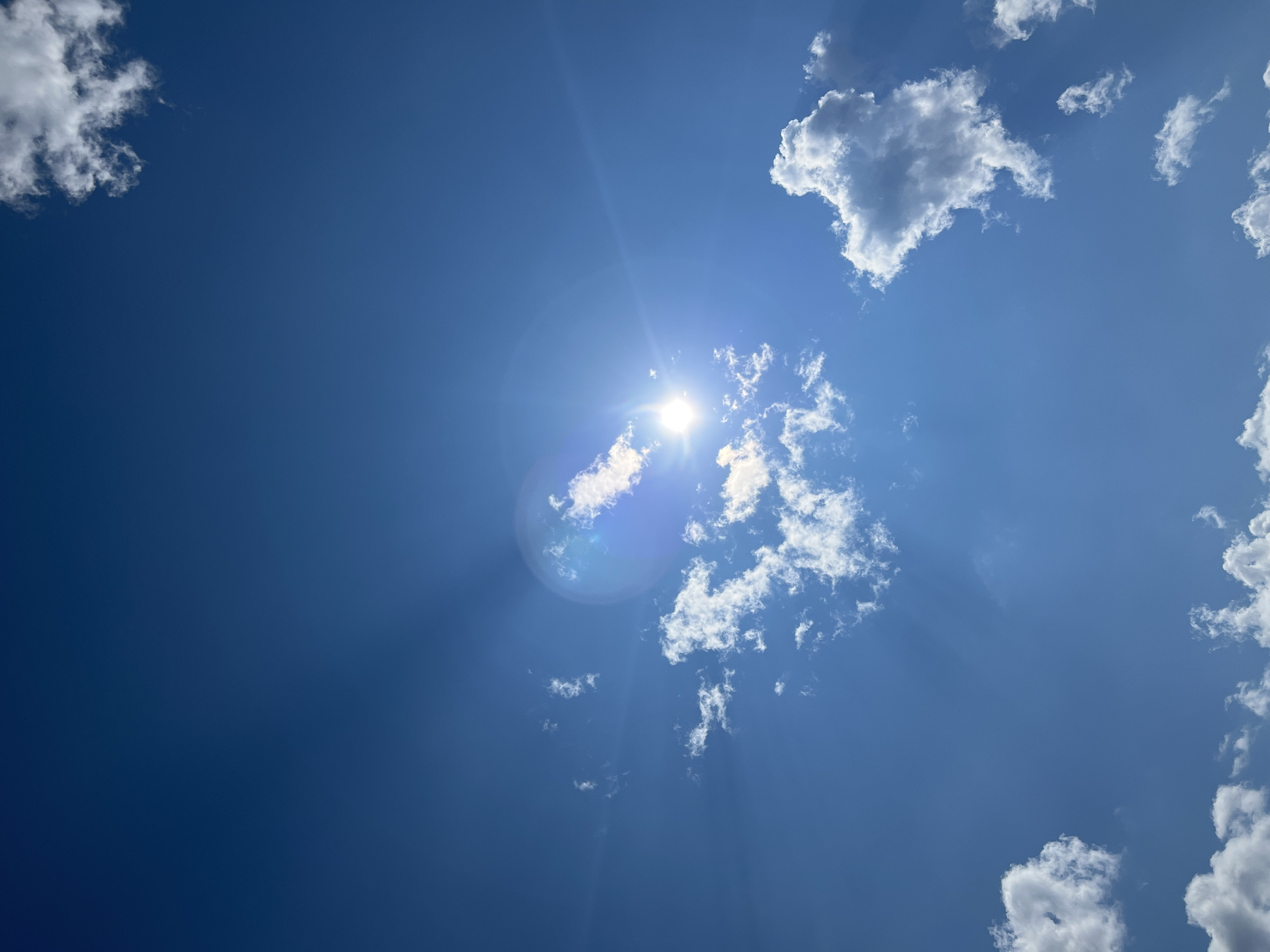 The image displays a clear blue sky with scattered white cumulus clouds. The sun is visible, creating lens flare and rays of light that emanate outwards. There are no people, subjects, or objects depicted in the scene, nor are any actions or interactions taking place. The environment is solely the sky. The provided location, Сосновка, Russia, is not visually represented in the image.