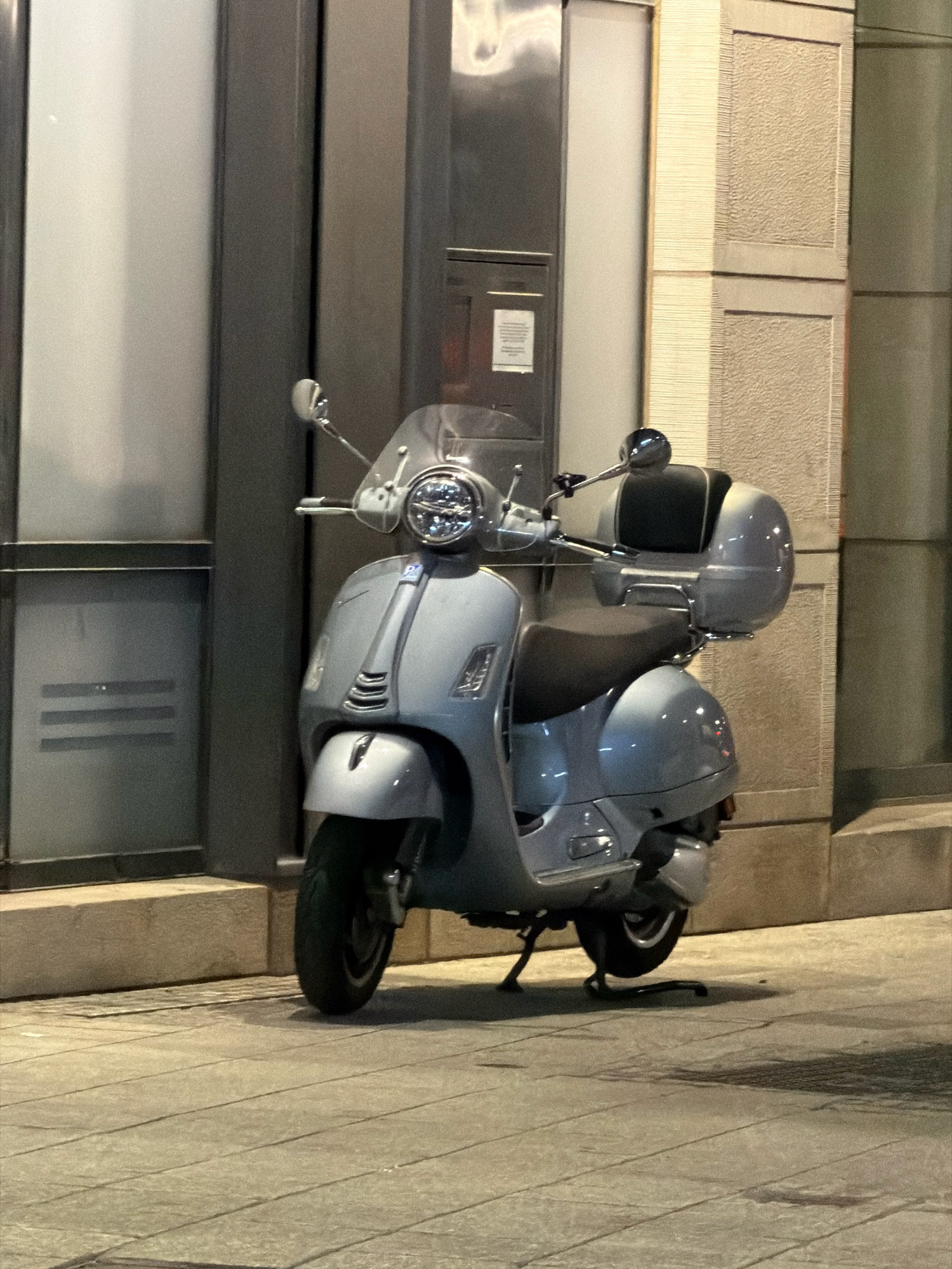A light blue Vespa scooter is parked on a sidewalk in front of a building. The scooter has a windscreen, a black seat, and a silver storage box on the back. The building appears to be modern, with glass and stone elements. The sidewalk is paved with large rectangular stones. The lighting suggests it is evening or night. There are no people visible in the image. The location is identified as Esch-sur-Alzette, Luxembourg.