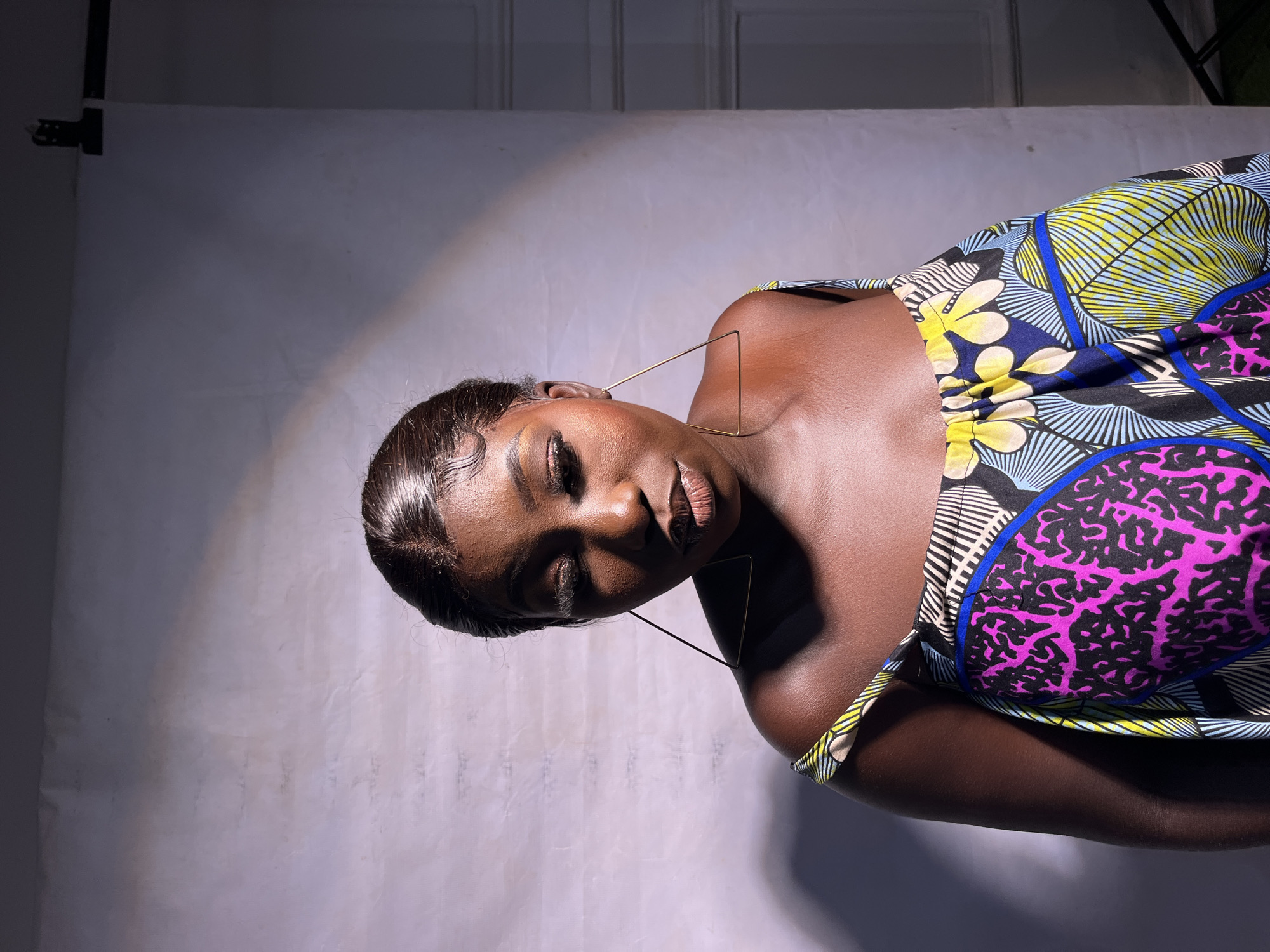 A portrait of a young Black woman with dark skin and dark hair styled in curls and a center part. She is wearing a vibrant, off-the-shoulder dress with a colorful African print. She has large, triangular gold earrings and wears dramatic false eyelashes and eye makeup. Her eyes are closed, and her lips are slightly parted. She is lit by a strong light source from the right, casting a shadow on the left side of her face and shoulder. The background is a plain white studio backdrop, with some subtle lighting effects creating a soft gradient. The setting is a photography studio in Jos, Nigeria.