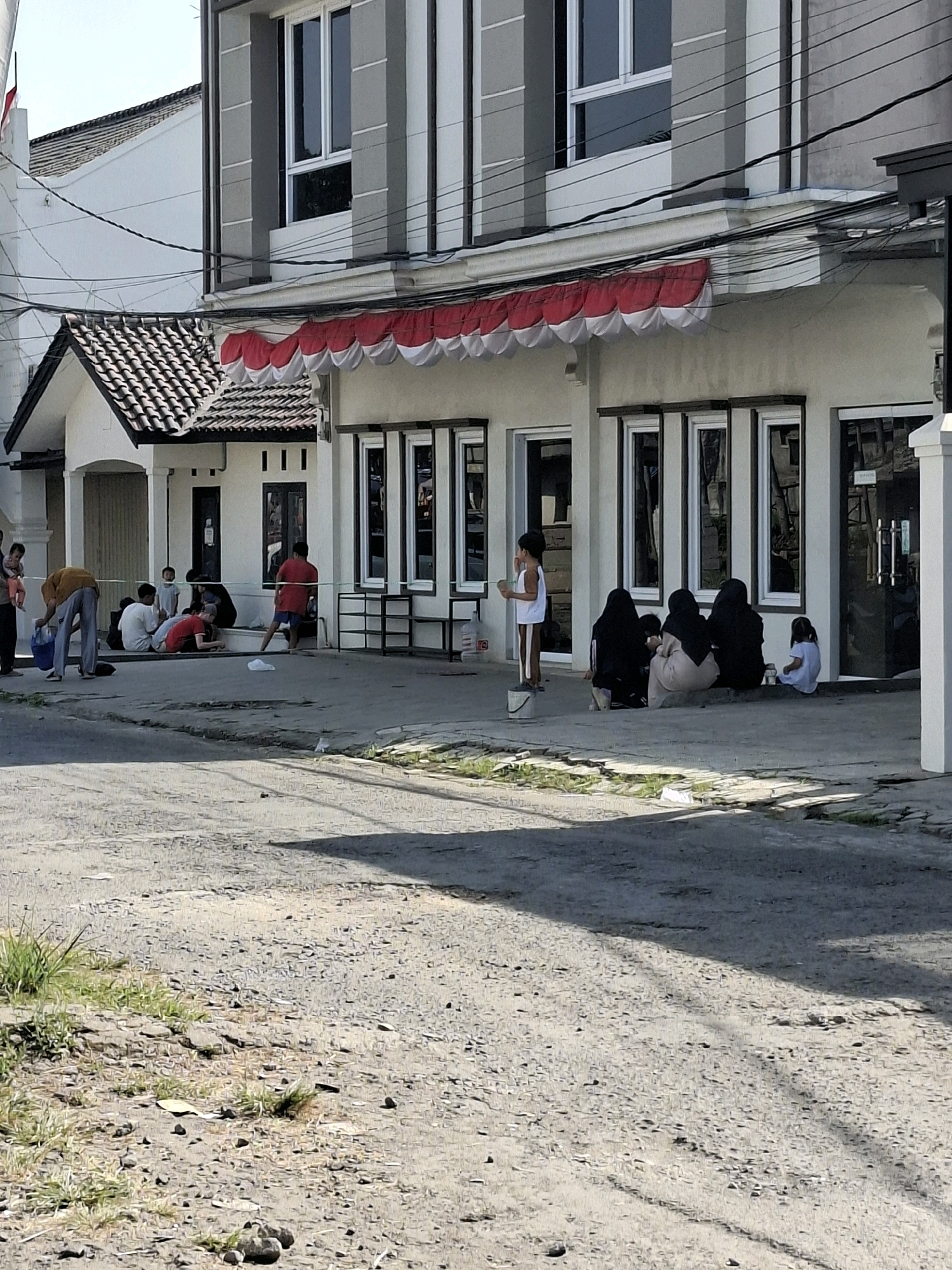 In the city of Karangtengah, Indonesia, a group of people are gathered on a concrete sidewalk in front of a modern building. The building features a white facade with dark-framed windows and a decorative red and white awning. To the left, a smaller building with a tiled roof is connected to the main structure. A few individuals are seated on the ground, while others stand. One person in an orange shirt is bending down, possibly interacting with something on the ground. Another person in a red shirt is standing near a green barrier. The ground is a mixture of paved concrete and dirt, with some sparse vegetation growing through cracks. Overhead, electrical wires crisscross the sky. The scene appears to be outdoors during the daytime.